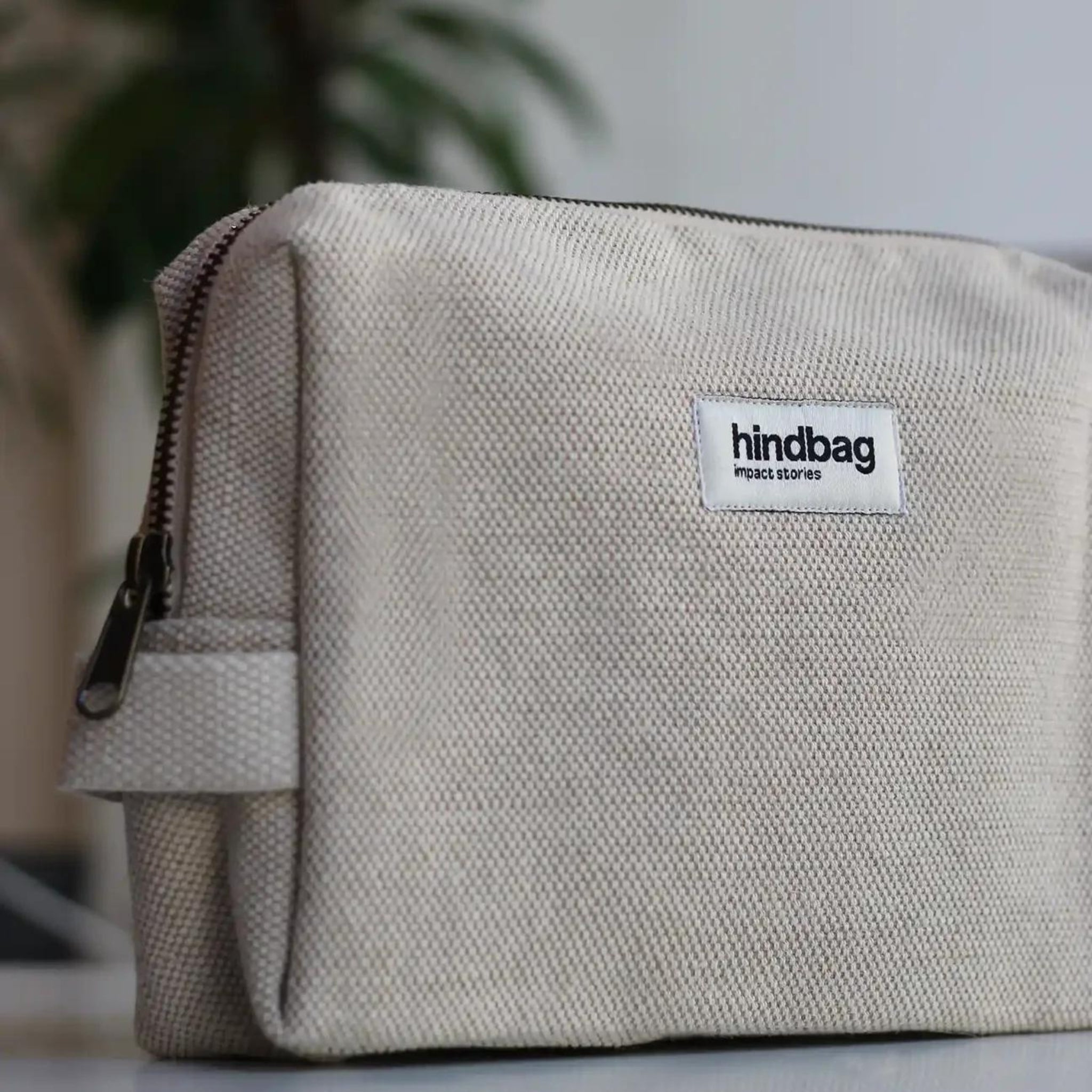 De Hindbag Leon Toilettas Jute van Hindbag, gemaakt van biologisch katoen met een rits en een klein label met impactverhalen, rust op een wit oppervlak met een vage groene plant op de achtergrond.