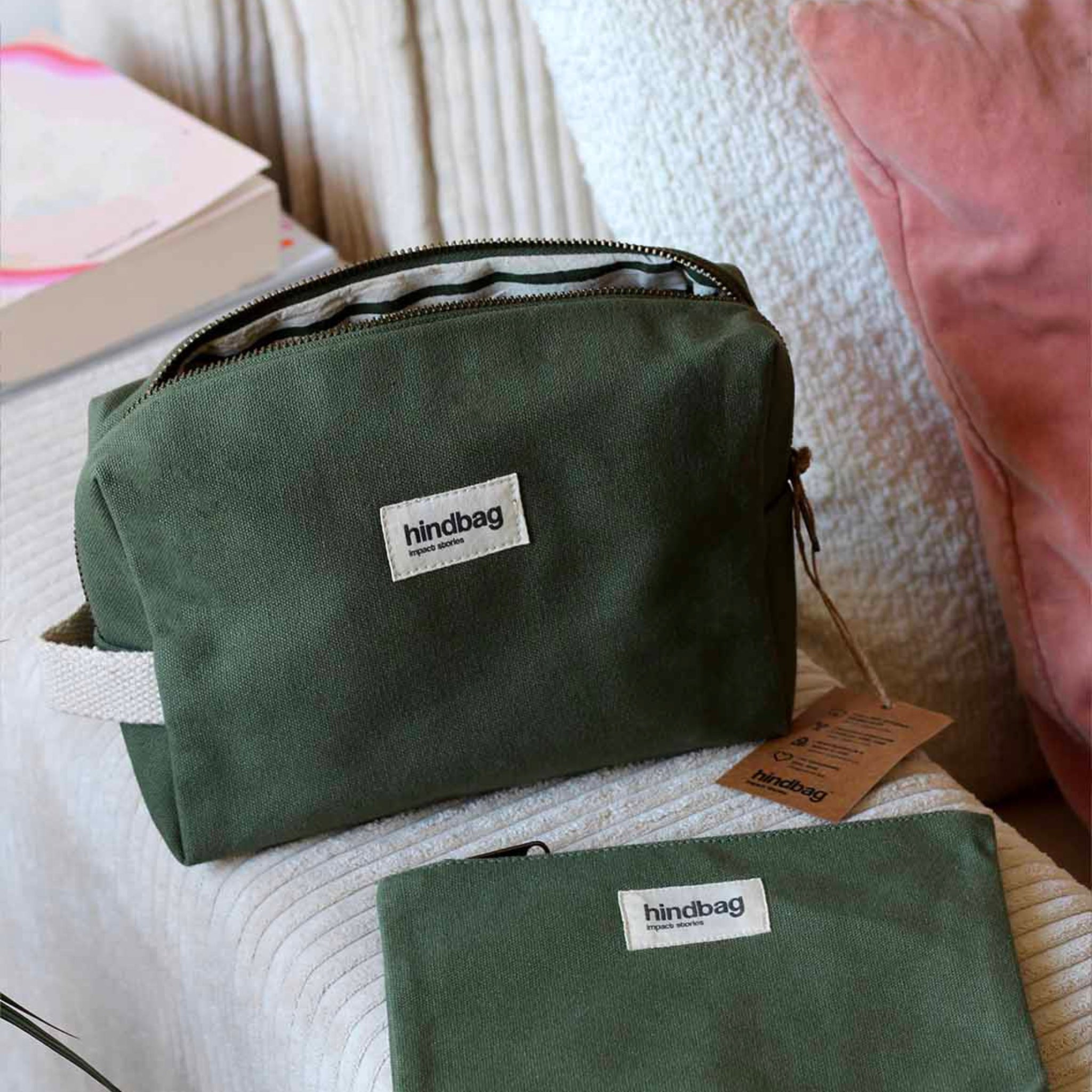 De Hindbag Leon Toilettas Olijfgroen van Hindbag, gemaakt van duurzaam organisch katoen met wit label, ligt op een beige ondergrond naast een bijpassend etui, roze kussen, boek en bruine tag.