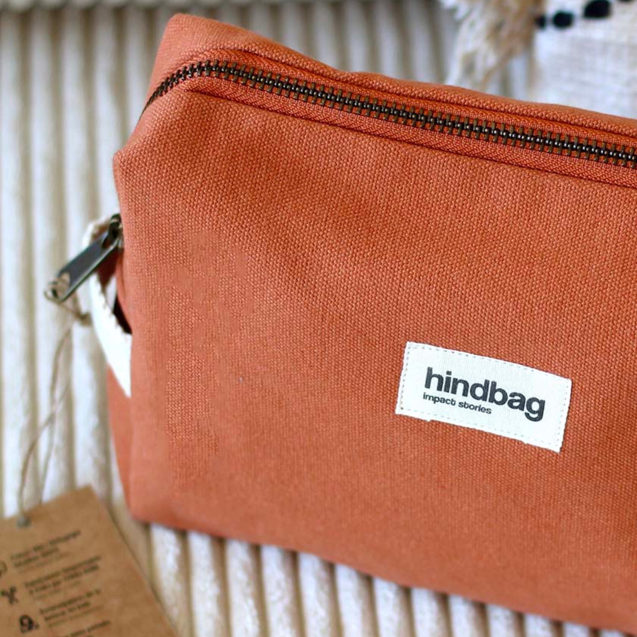 De Hindbag Leon Toilettas Siena, gemaakt van biologisch katoen in roestoranje, heeft een zwarte ritssluiting en een wit label met de tekst "Hindbag impact stories". Een bruin label is gedeeltelijk zichtbaar aan de linkerkant als het rust op een beige oppervlak met textuur.