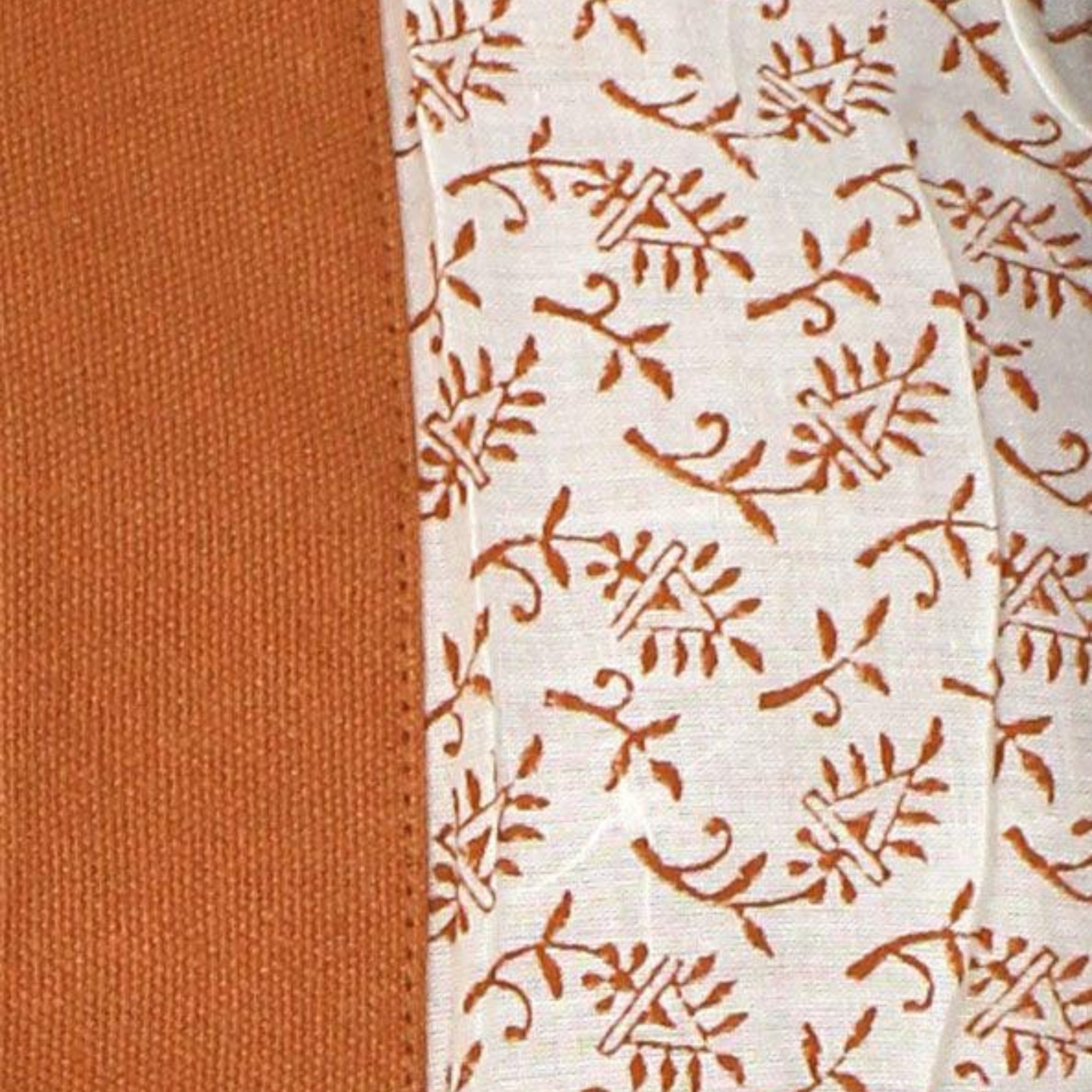 Close-up van twee stoffen uit de Hindbag Leon Toilettas Siena van Hindbag: een effen roestoranje, de andere wit met een roestoranje bloemenrankenpatroon, beide van organisch katoen. Hun texturen zorgen voor een opvallend contrast.