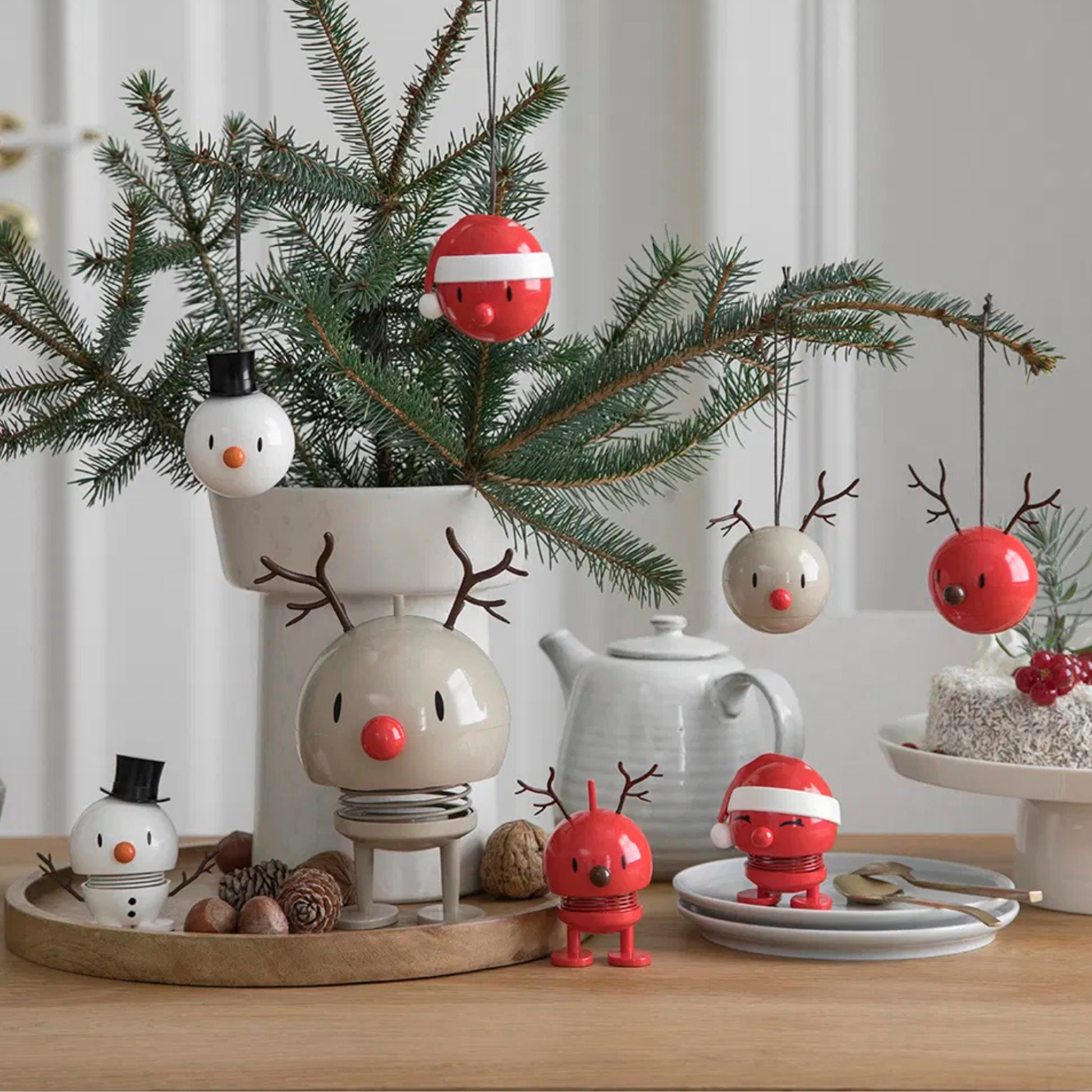 Een feestelijke tafel met kleine kerstdecoratie, zoals de Sneeuwman Bumble S Wit van Hoptimist, genesteld tussen dennentakken, een theepot, cake en borden creëert een vrolijk kersttafereel dat perfect is voor elk cadeautje-moment.