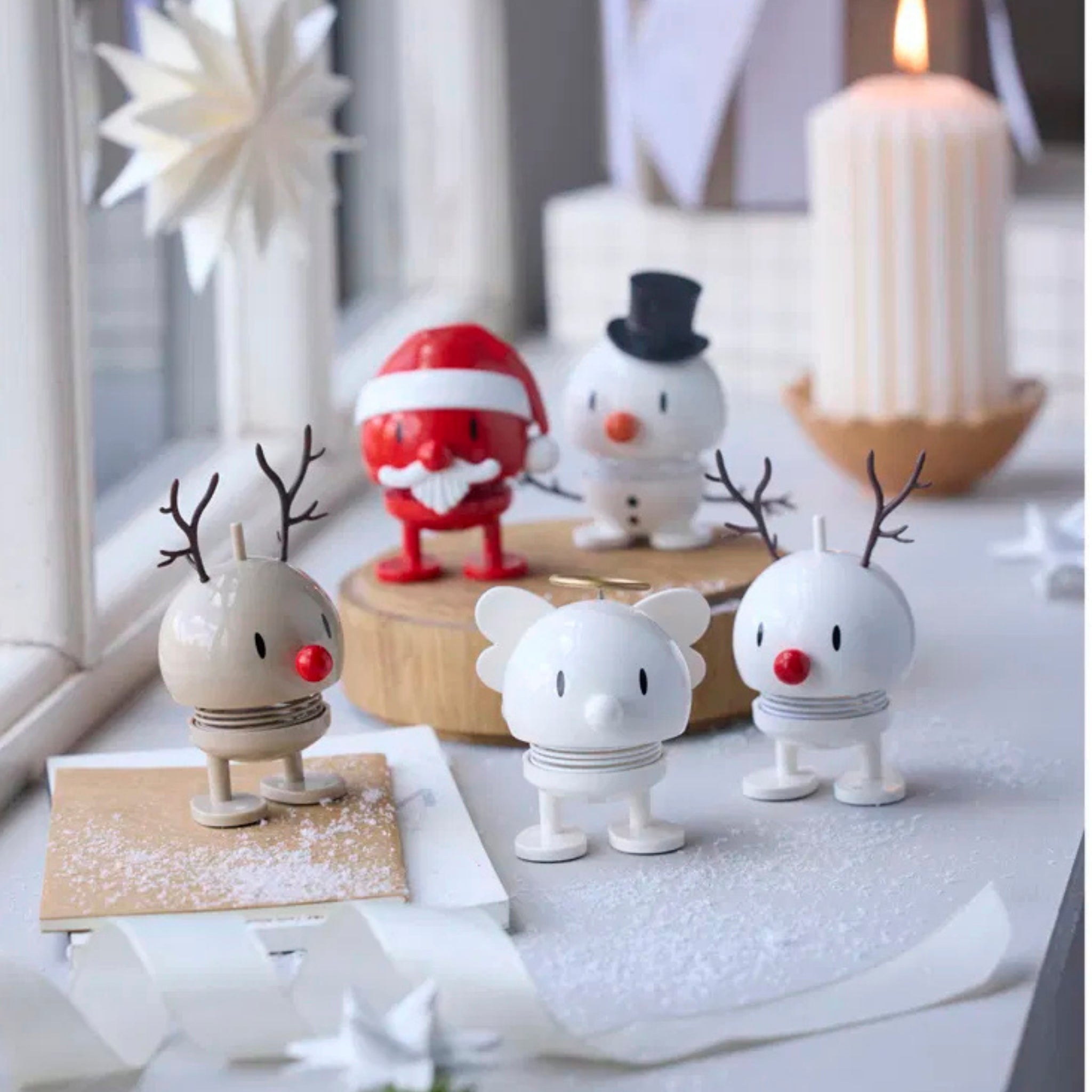 Vier kleine Hoptimist Engel Bumble S Wit beeldjes, geïnspireerd op het Deense ontwerp van Gustav Ehrenreich, staan op een besneeuwd oppervlak naast de Kerstman en een sneeuwpop verlicht door kaarslicht en sterversieringen bij het raam.