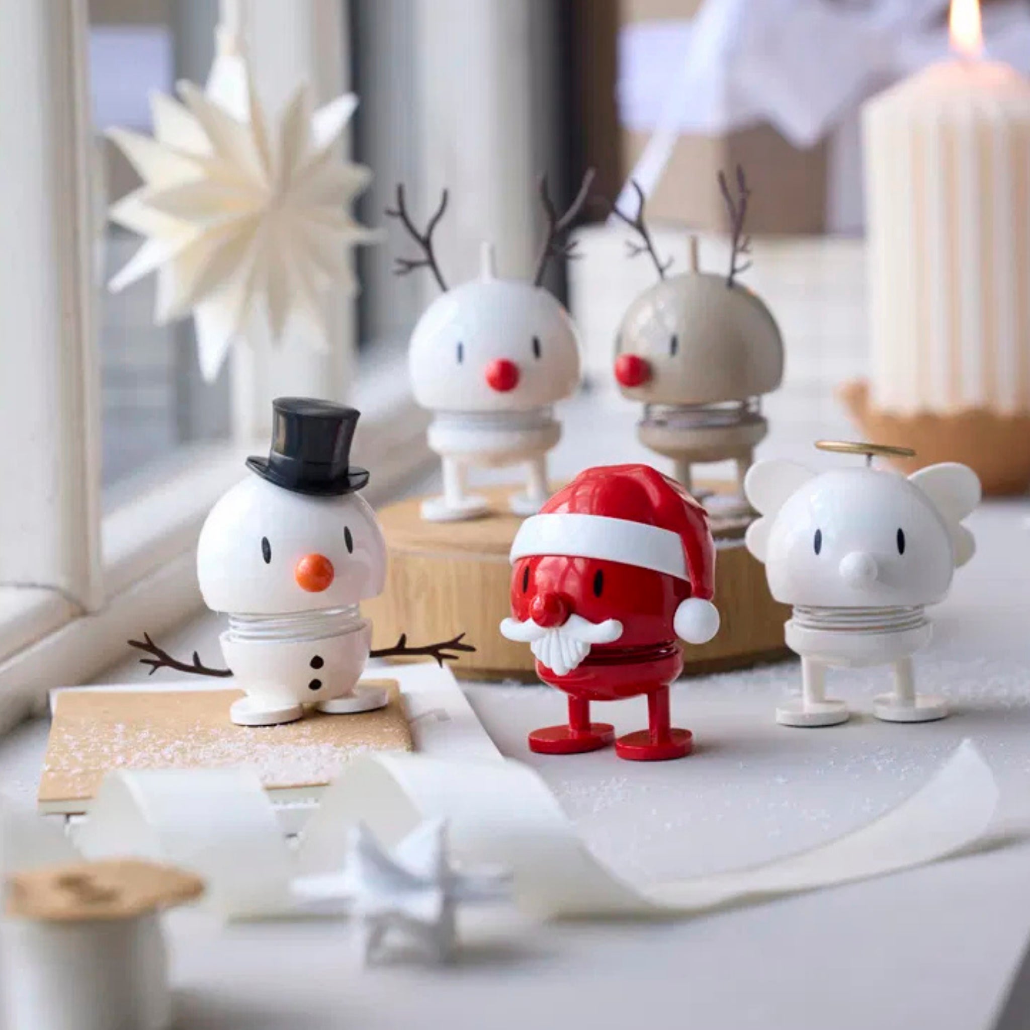 Vier kleine, ronde Hoptimist Engel Bumble S Wit beeldjes - een sneeuwpop, Kerstman, rendier en ijsbeer - staan op een vensterbank. Op de zacht verlichte achtergrond staan decoratieve voorwerpen, een witte kaars en een papieren sterornament.