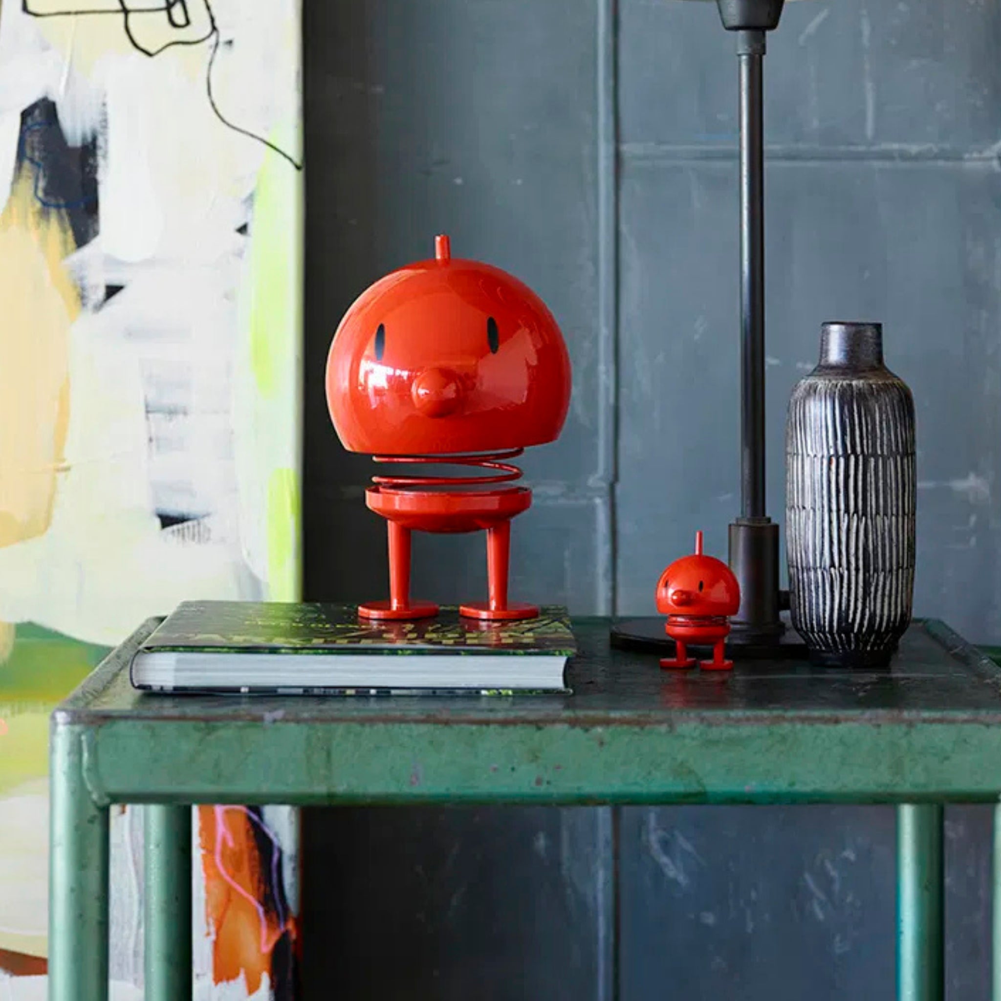 Op een groene tafel staan twee Hoptimist Bumble S Rood veerspeeltjes, een grote en een kleine van Hoptimist, met op de achtergrond een boek, een grijze geribbelde vaas, een zwarte lamp, abstracte kunst en een grijze betegelde muur met Deens ontwerp.