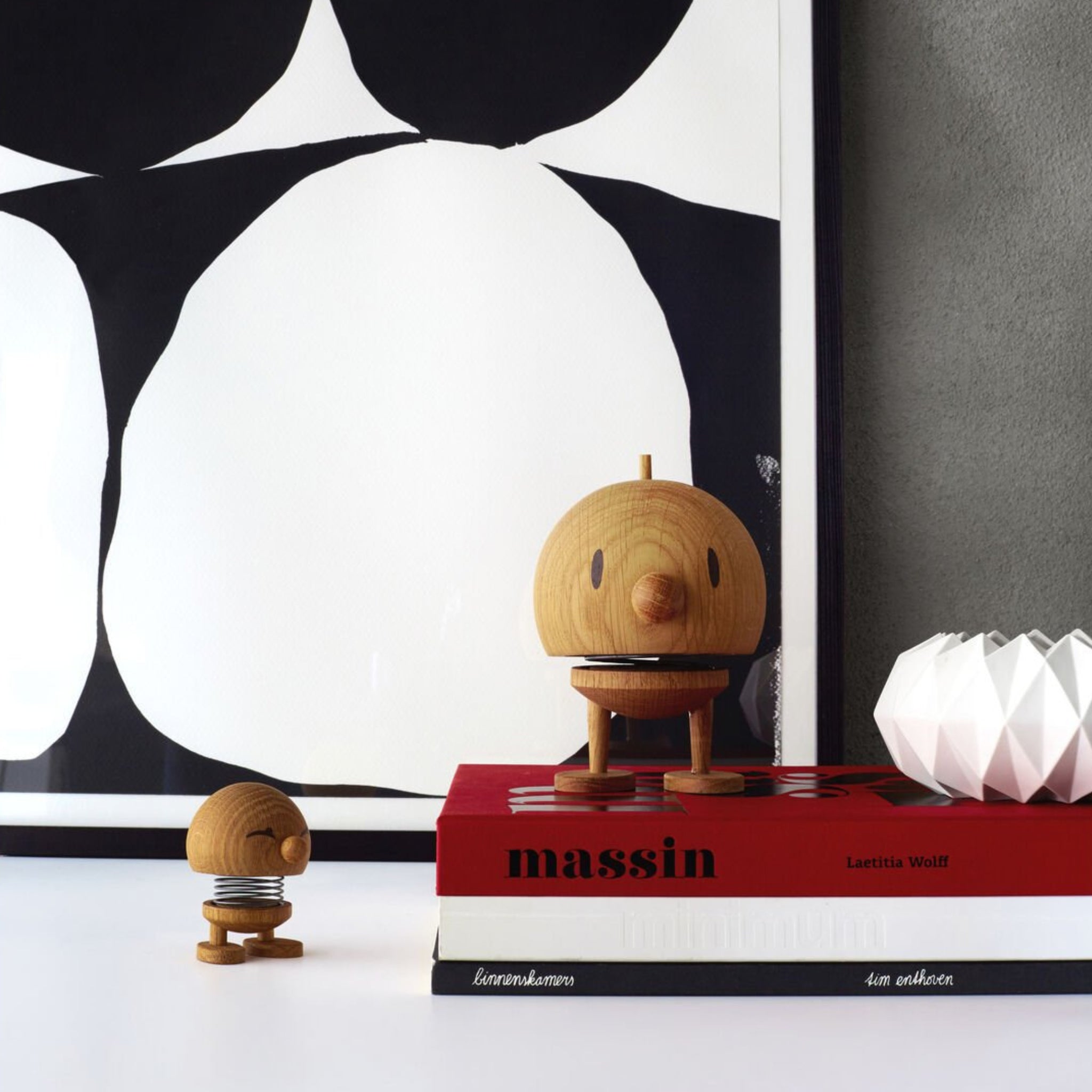 Twee Hoptimist Hout Bumble S Eik beeldjes staan op een wit oppervlak; één is geplaatst bovenop gestapelde rode en zwarte boeken, naast een witte geometrische vaas met abstracte zwart-witte kunst op de achtergrond.