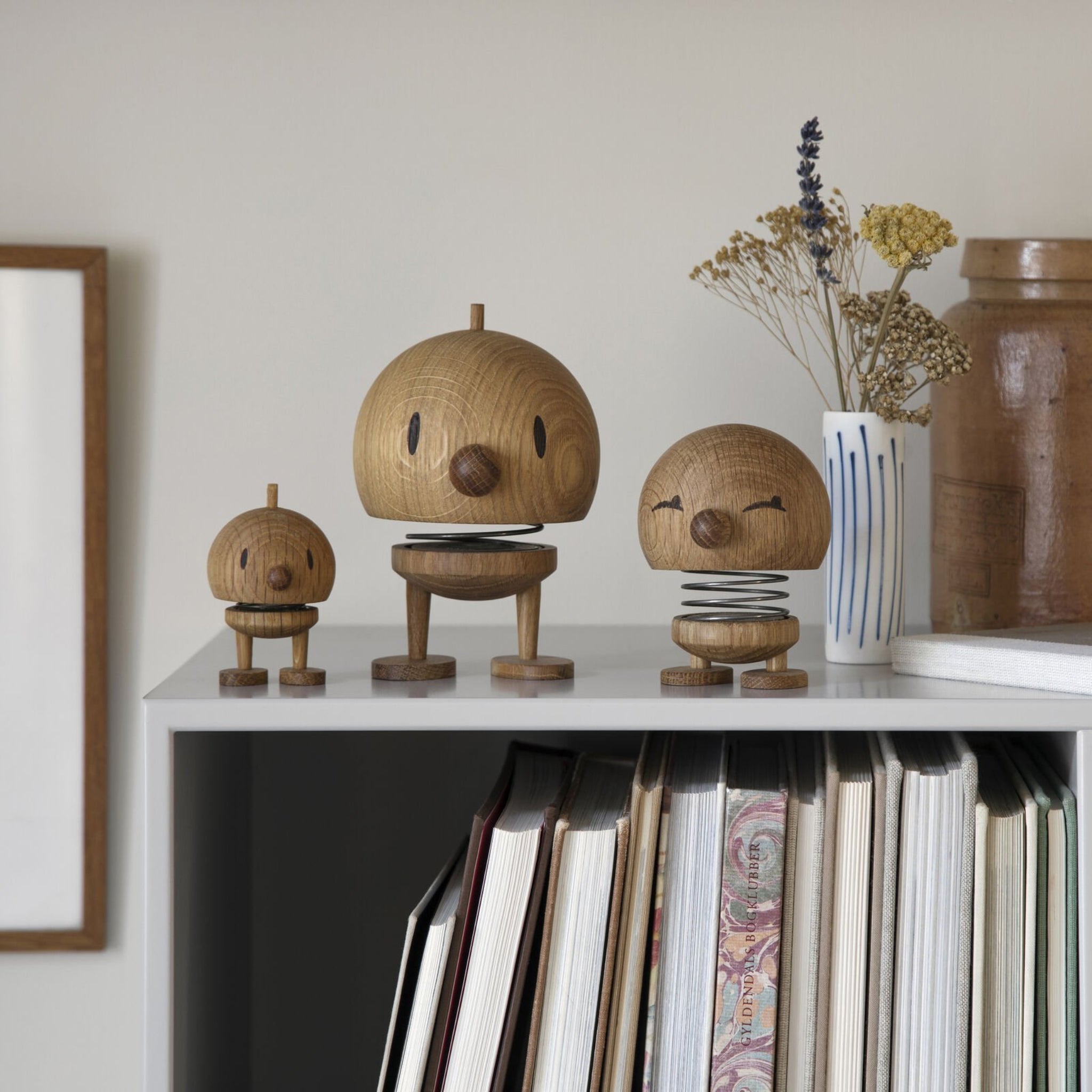 Drie houten Hoptimist Hout Bumble S Eik beeldjes met ronde hoofden en eenvoudige gezichten staan op een boekenplank vol boeken, terwijl een witte vaas met gedroogde bloemen en een bruine keramieken pot de Deens design uitstraling versterken.