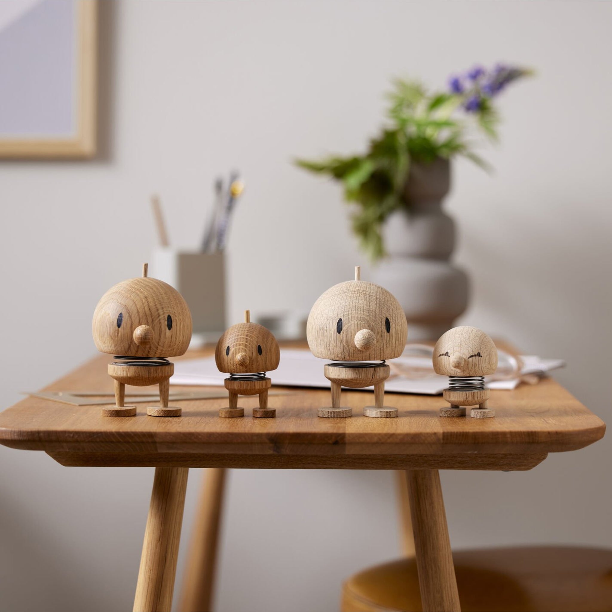Vier Hoptimist Hout Bumble S Eik beeldjes met ronde hoofden en eenvoudige kenmerken staan op een houten tafel, omringd door Deens designelementen zoals kantoorbenodigdheden, een bloemenvaas en vaag decor op de achtergrond.