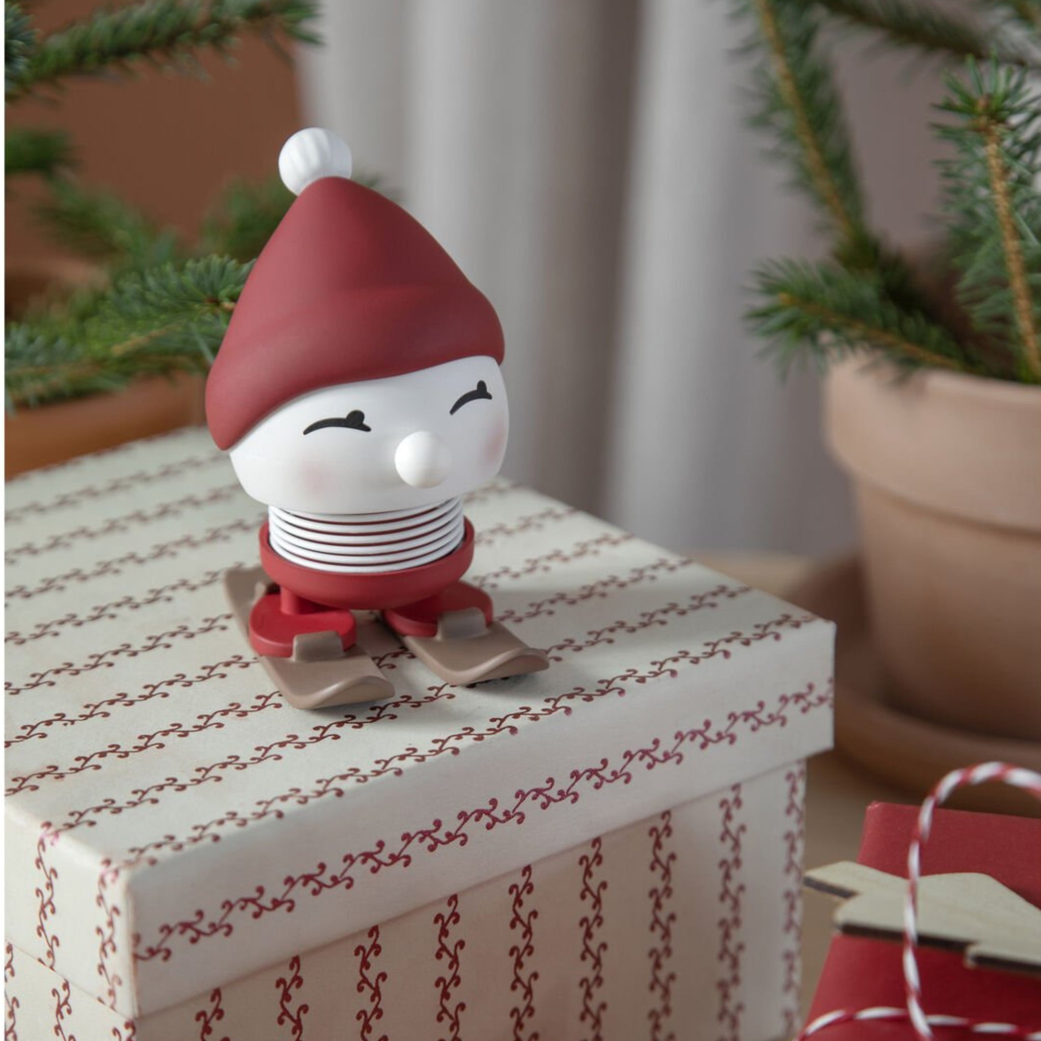 De Hoptimist Ski Bimble S Berry van Hoptimist - een klein, lachend beeldje met een rode muts en ski's - zit bovenop een geschenkdoos met een rood patroon. Het is een origineel geschenk en een perfecte winterdecoratie met dennentakken op de achtergrond.