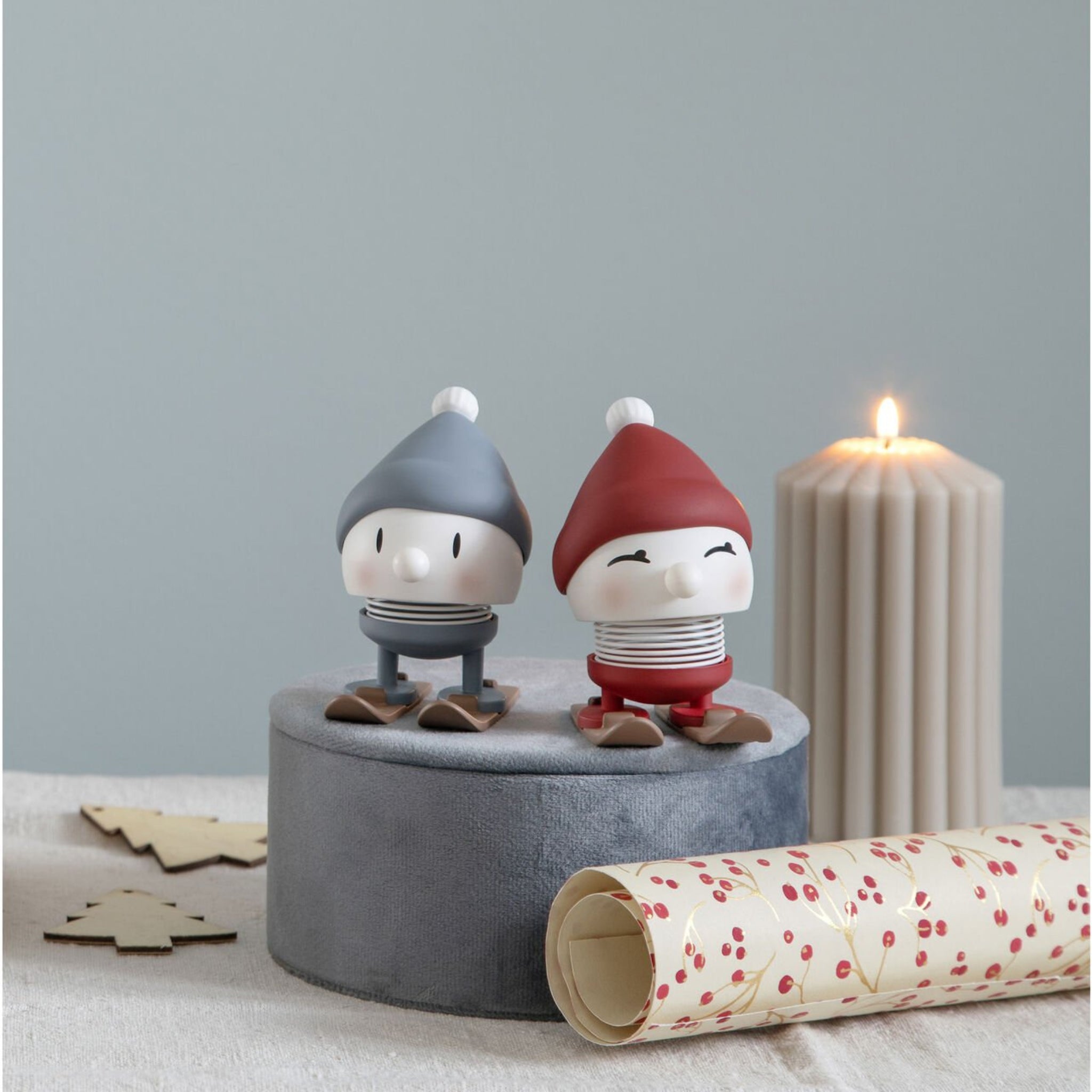 De Hoptimist Ski Bimble S Berry van Hoptimist heeft twee kleine figuurtjes met wintermutsen, een ronde grijze voet, een brandende beige kaars, houten bomen en feestelijk inpakpapier met rode bessen op lichtblauw.