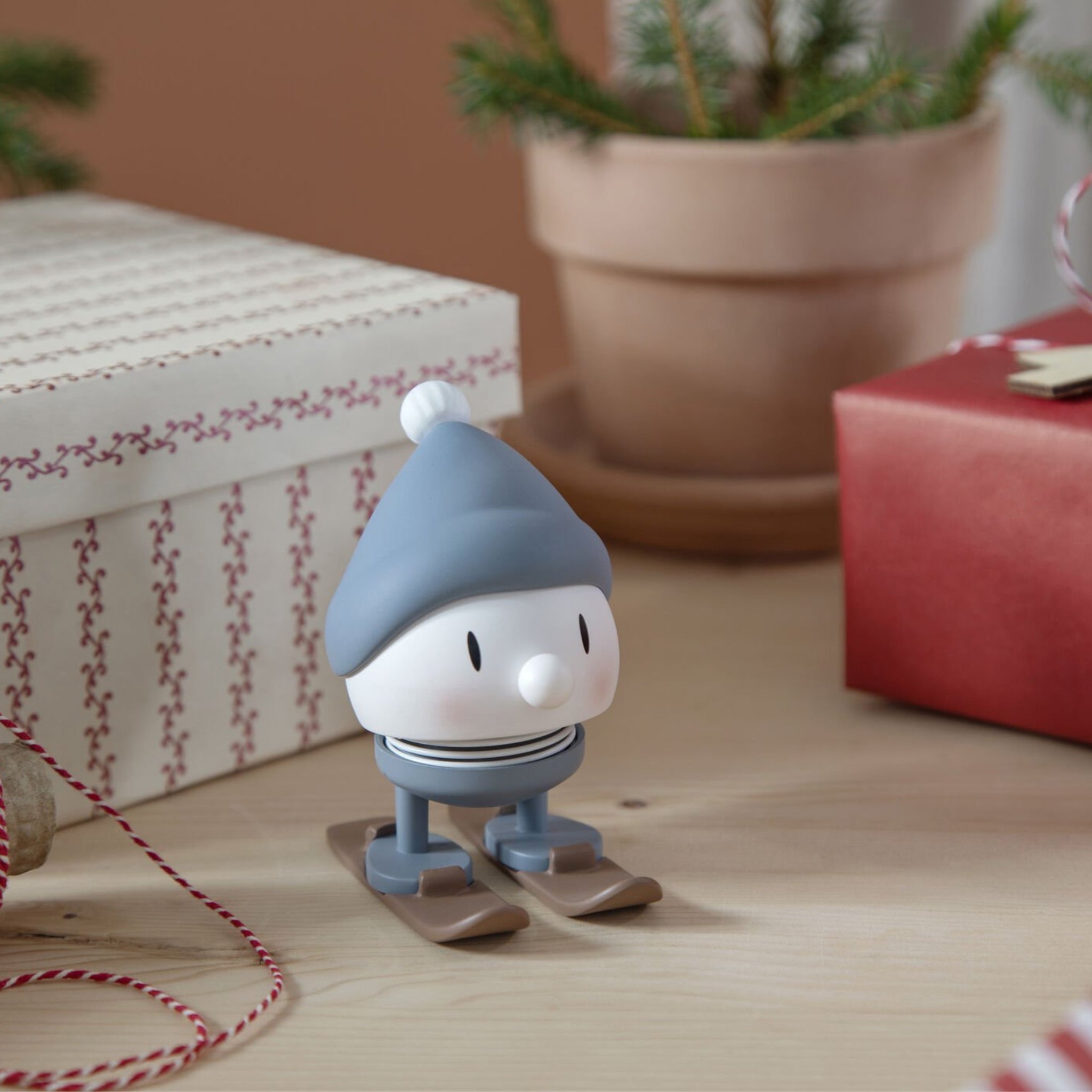 Het Hoptimist Ski Bumble S Sky beeldje, met een blauwe muts en sjaal, staat op ski's tussen ingepakte cadeautjes - een origineel cadeau en perfecte winterse decoratie met rood-wit garen en een potplant. Merk: Hoptimist.