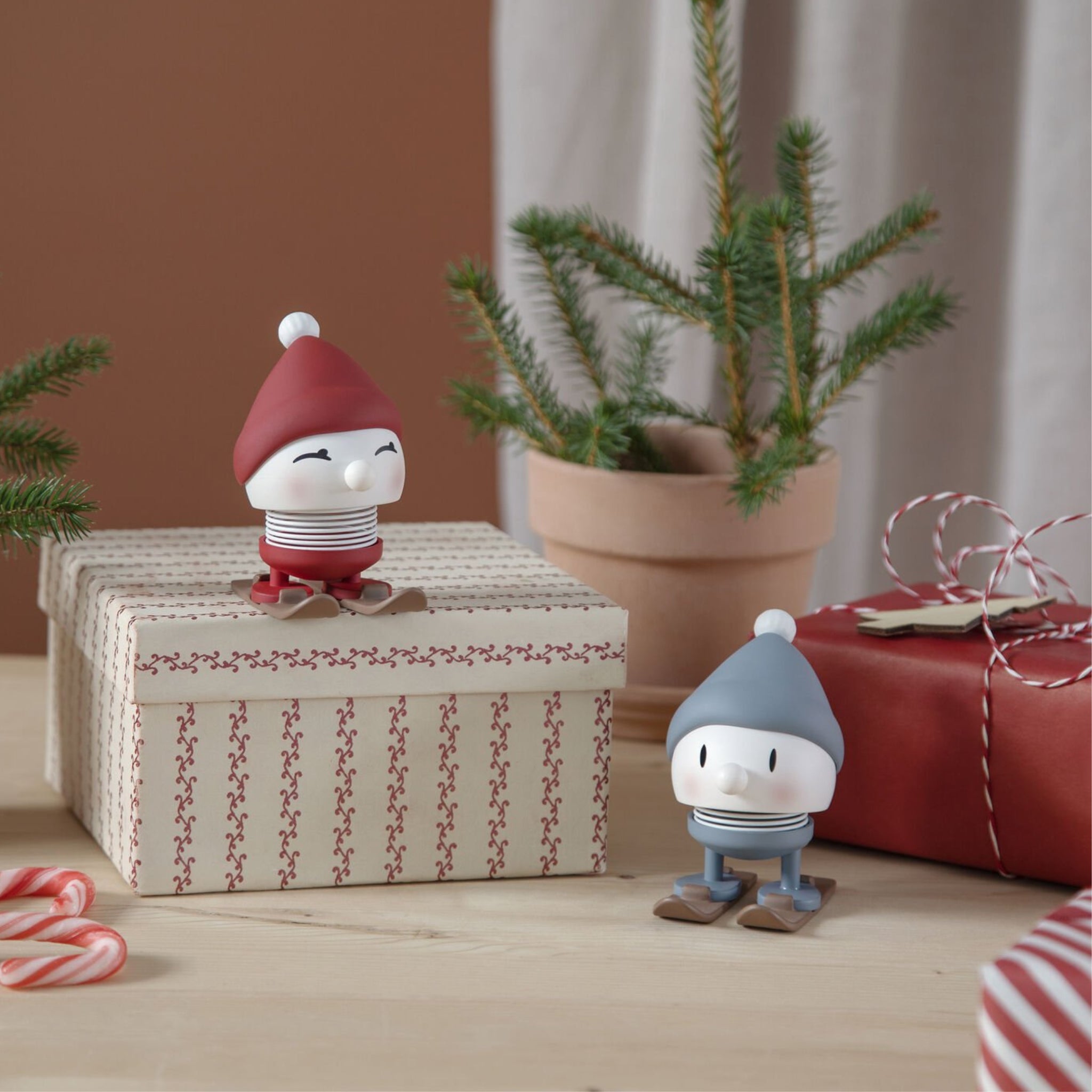 Twee beeldjes van Hoptimist Ski Bumble S Sky, met wintermutsen en sjaals, zitten op een geschenkdoos en tafel. Dit gezellige kersttafereel bevat een dennenboom in pot, zuurstokken, feestelijk koord en een origineel cadeau voor geliefden.