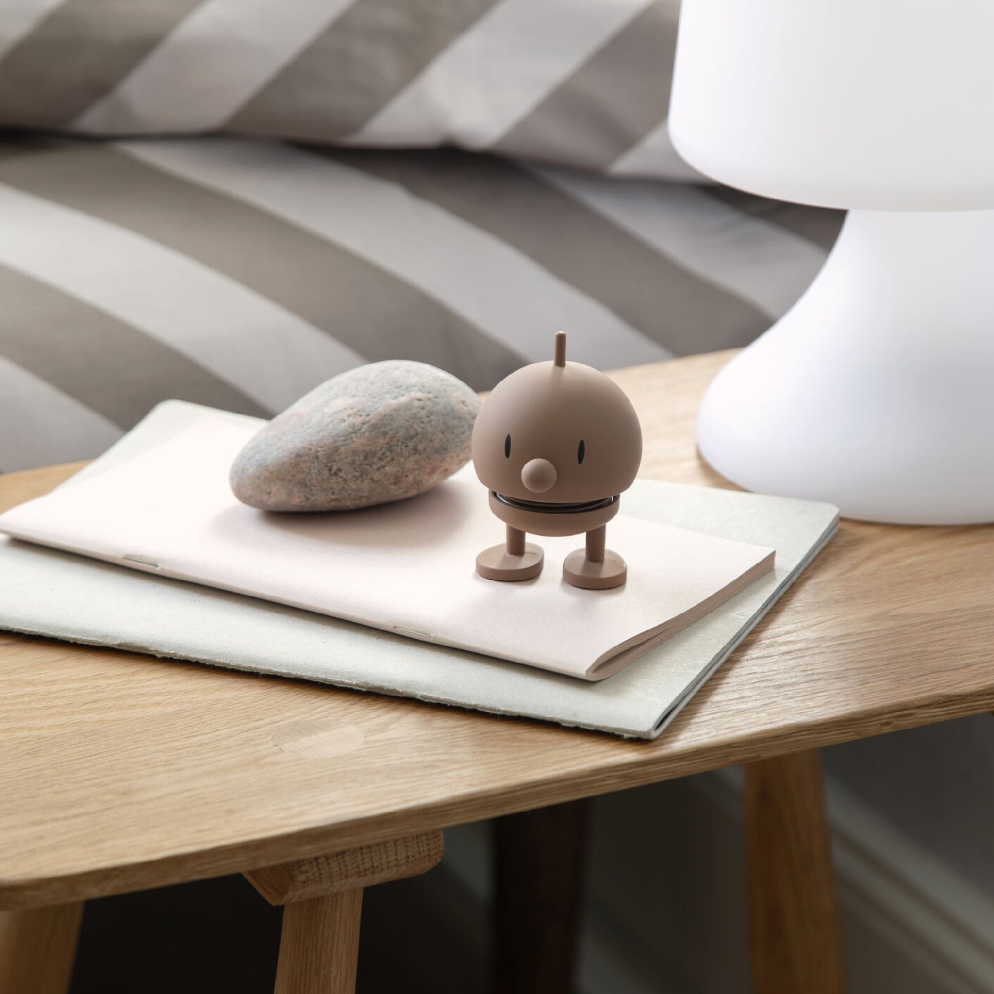 De Hoptimist Soft Bumble S Chocolate van Hoptimist - een klein bruin speelgoedfiguurtje - staat op een gesloten notitieblokje naast een gladde grijze steen, allemaal gerangschikt op een houten nachtkastje naast een witte lamp voor speelse Deense design charme.