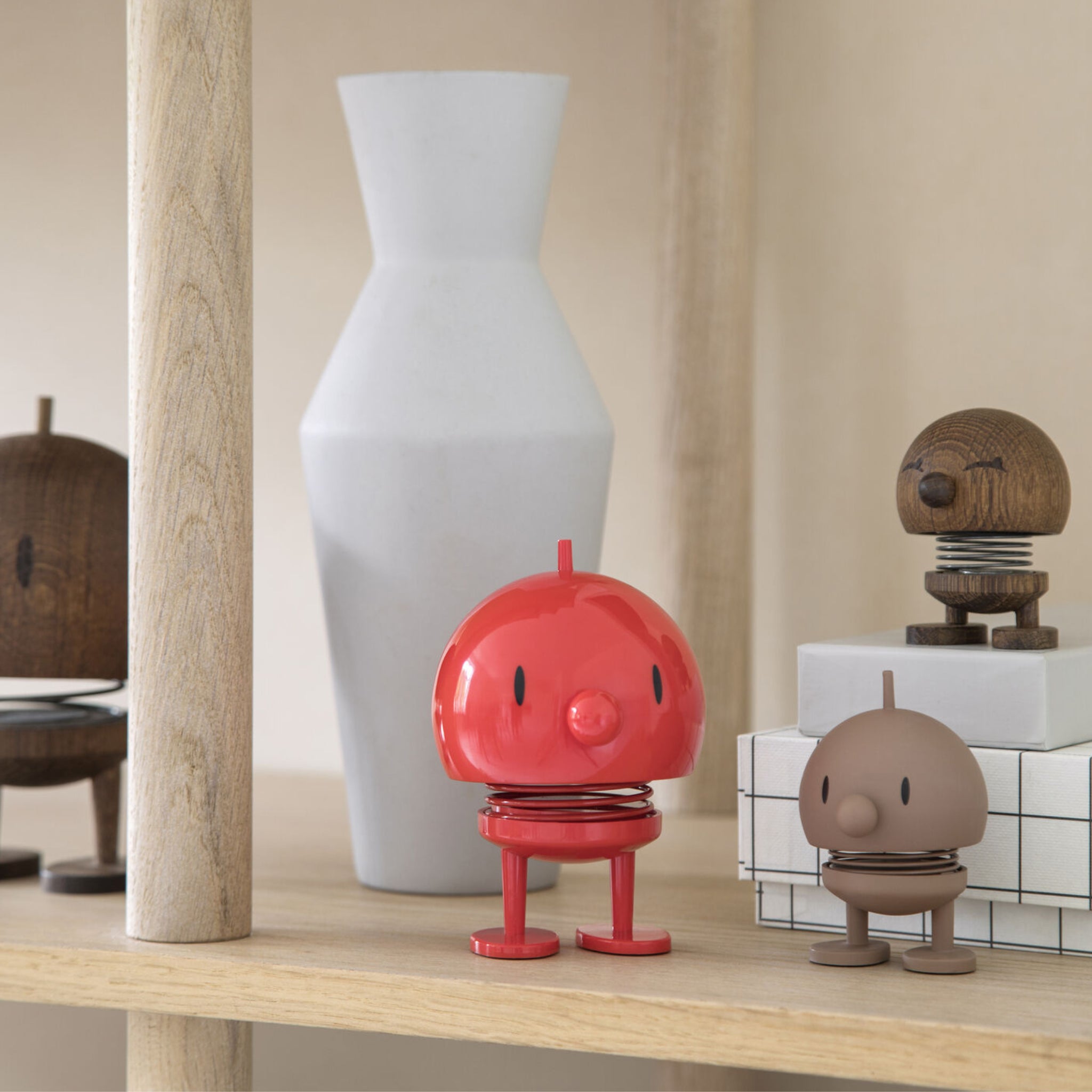 Een close-up van een schap toont het Hoptimist Soft Bumble S Chocolade figuurtje van Hoptimist, een klassiek Deens ontwerp in chocoladebruin, naast een witte geometrische vaas en gestapelde witte dozen.