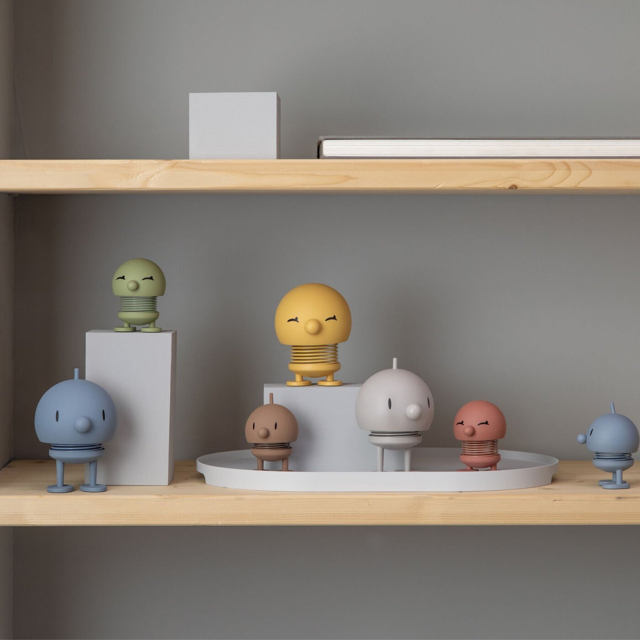 De Hoptimist Soft Bumble S Chocolade van Hoptimist, met zijn speelse veerlichaam en vrolijke gezicht, voegt een vleugje Deense design charme toe aan een minimalistische kamer wanneer hij wordt tentoongesteld op een houten plank tegen een grijze muur.