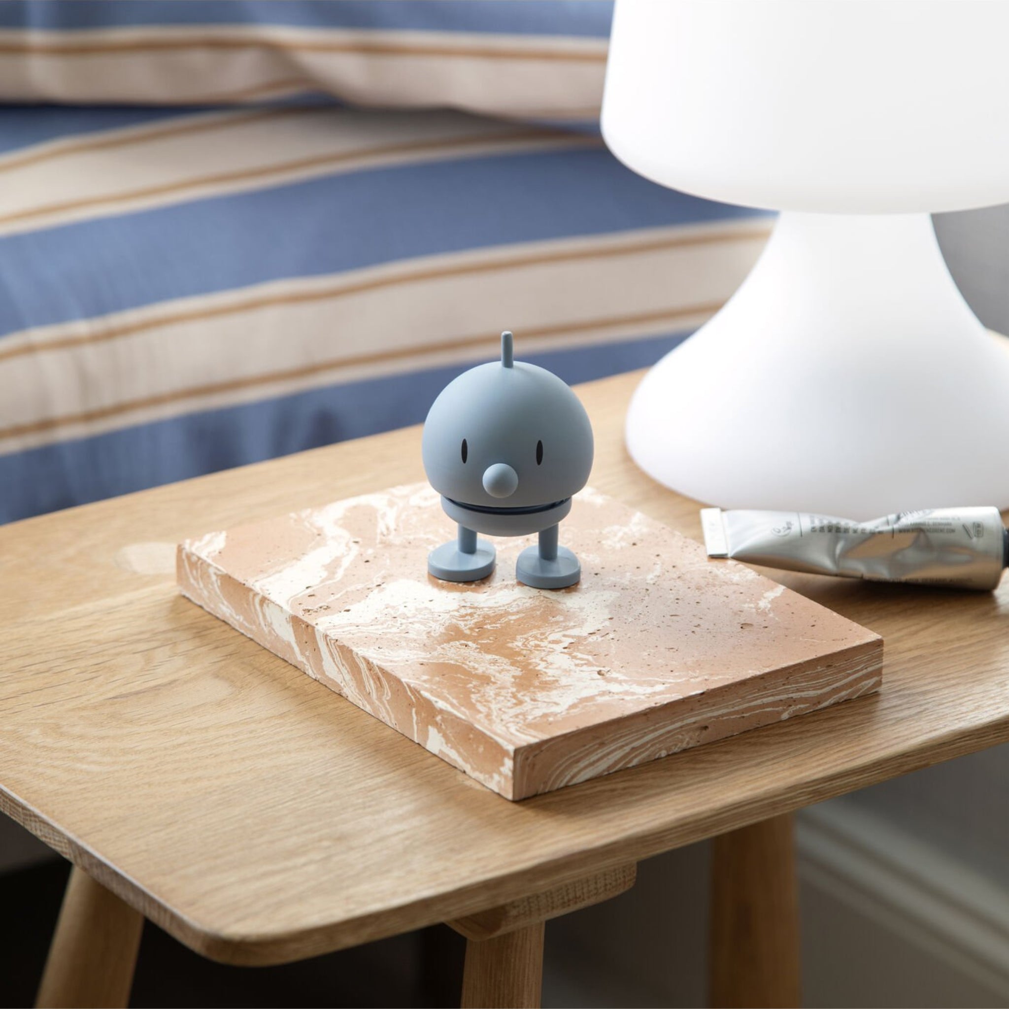 De Hoptimist Soft Bumble S Sky, een klein blauw figuurtje van Hoptimist, staat op een beige gemarmerd notitieboekje op een houten nachtkastje naast een witte lamp en crème, met een gestreept bed op de achtergrond - echt Deens design.