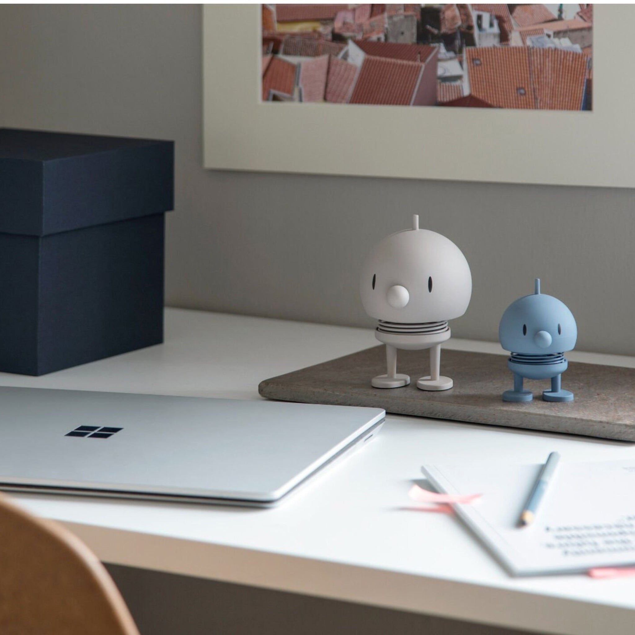 Een minimalistisch bureau met een gesloten laptop, twee Hoptimist Soft Bumble S Sky beeldjes van Hoptimist - een echo van Deens design - plus potloden, papieren, een blauwe doos en een ingelijste foto van het dak aan de muur.