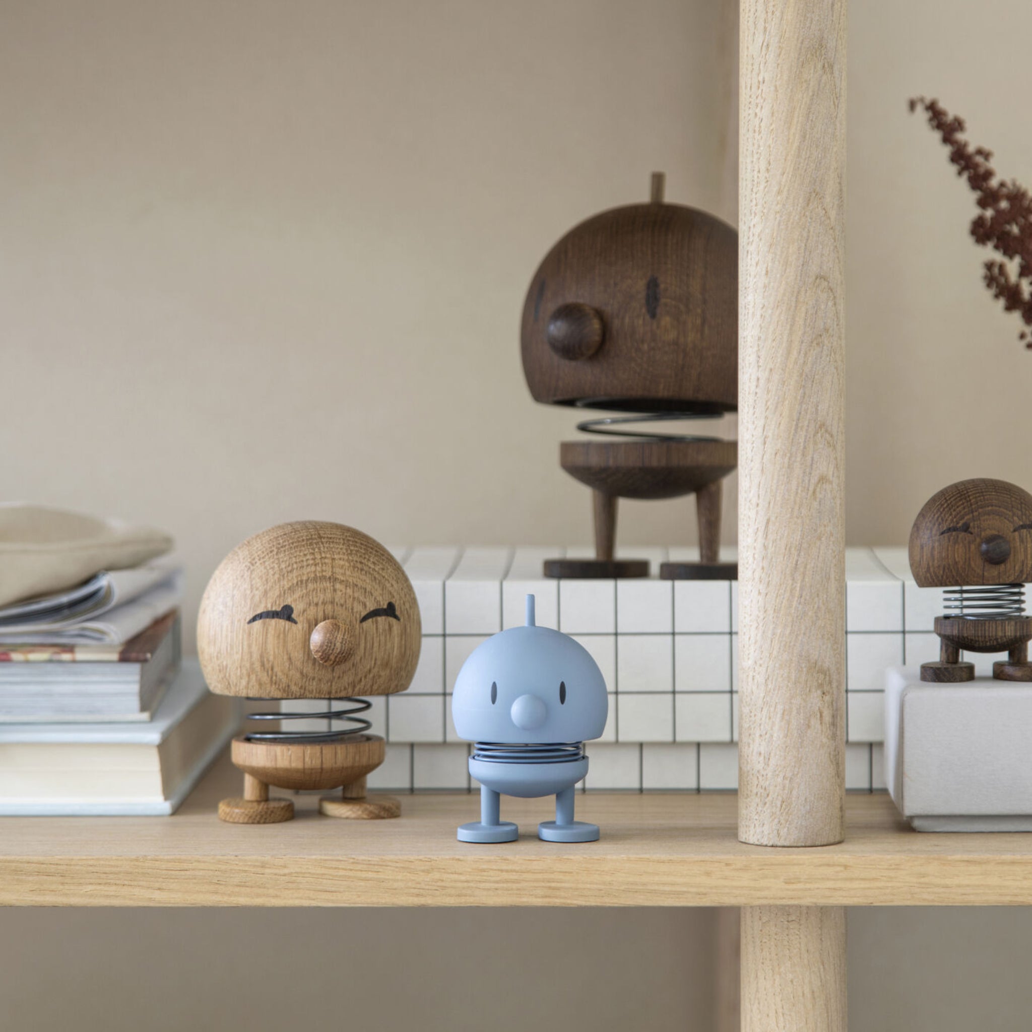Drie houten Hoptimist beeldjes en één Hoptimist Soft Bumble S Sky van Hoptimist, allemaal met ronde hoofden en veerlichamen, staan op een houten plank met boeken en neutrale decoraties in een minimalistische Deense designomgeving.