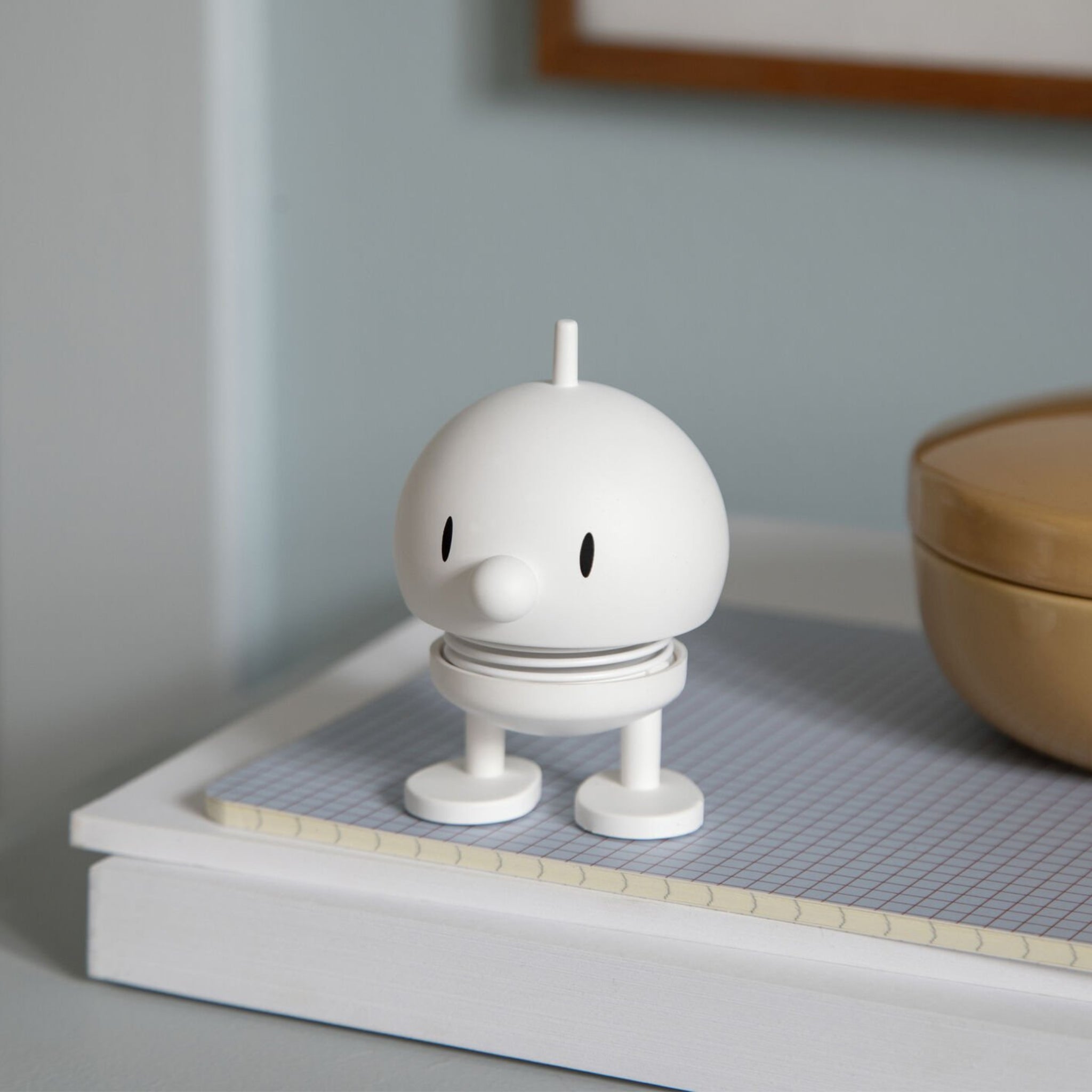 De Hoptimist Soft Bumble S Wit, een klein, wit beeldje van Hoptimist met een groot hoofd en ronde neus, staat op een dienblad met rasterpatroon naast een beige bakje tegen een lichtblauwe achtergrond, geïnspireerd op Deens design.