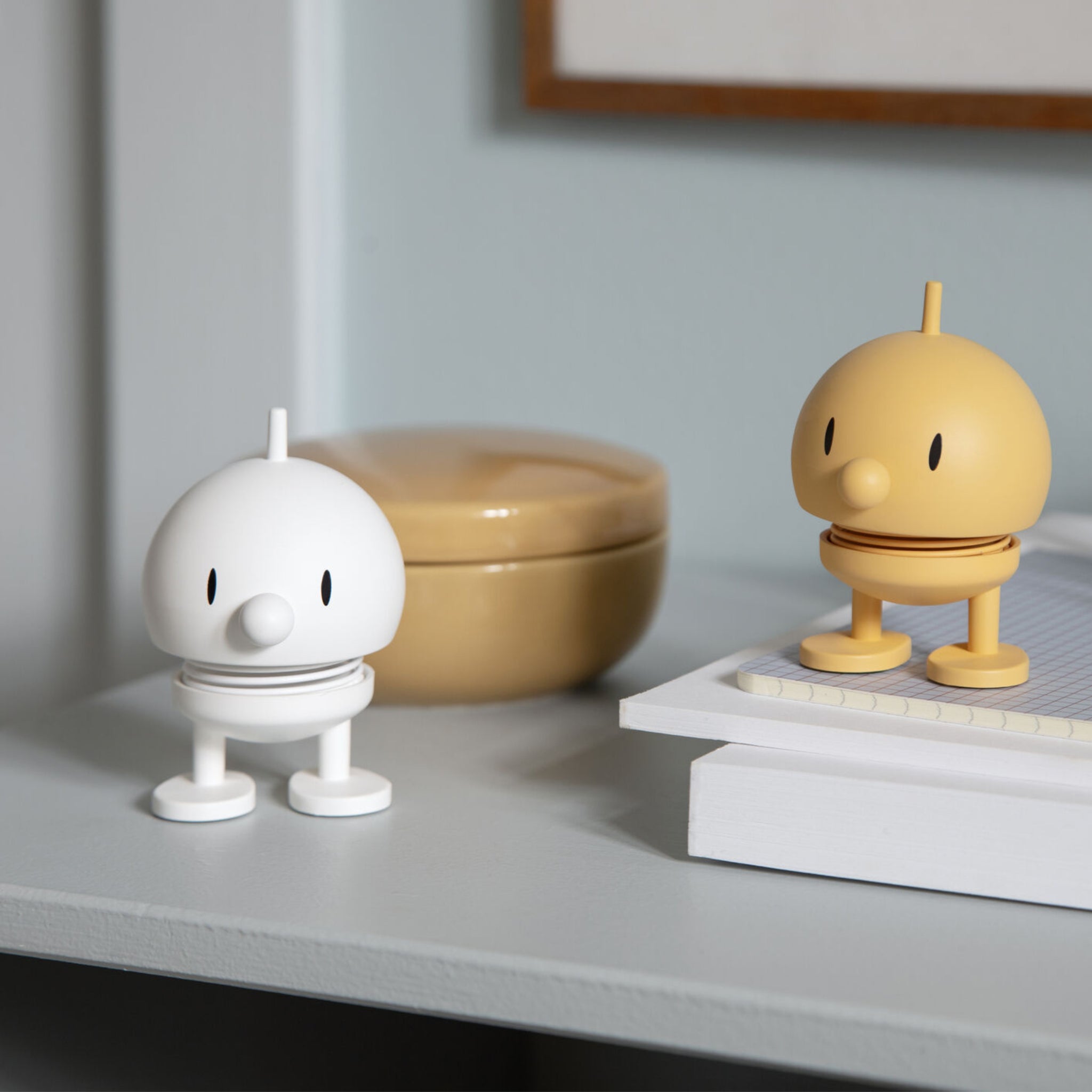 Een wit Hoptimist Soft Bumble S beeldje van Hoptimist staat op een plank naast een gesloten notitieboekje en een beige container en voegt speels Deens design toe aan de scène tegen een lichte muur met ingelijste foto.