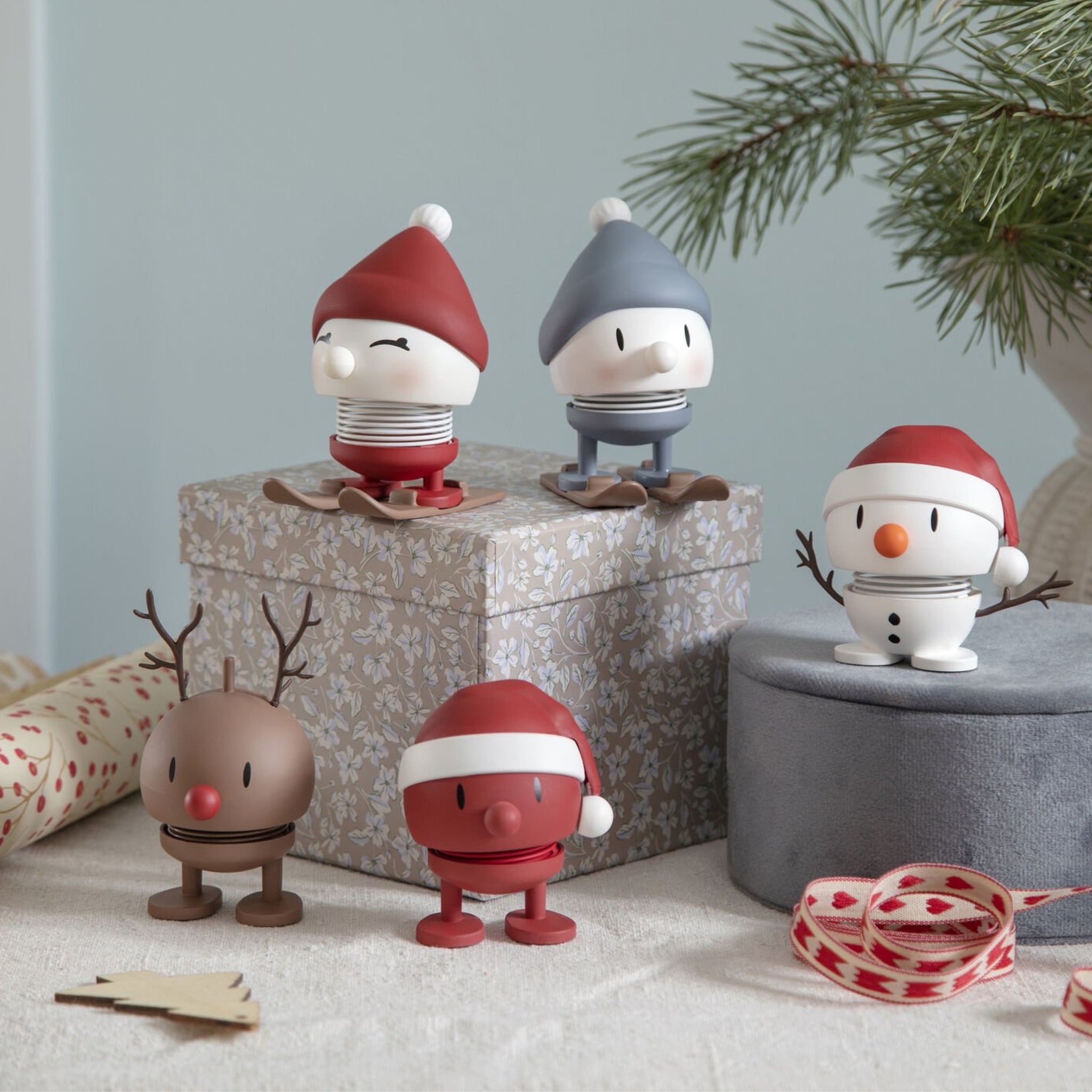 Vijf kleine, ronde Hoptimist-figuurtjes met lentepootjes - waaronder de Zachte Kerstman Hommel S Rood - staan tussen geschenkdozen, feestelijke linten en sparrentakken voor een vrolijk Deens tintje dit seizoen.