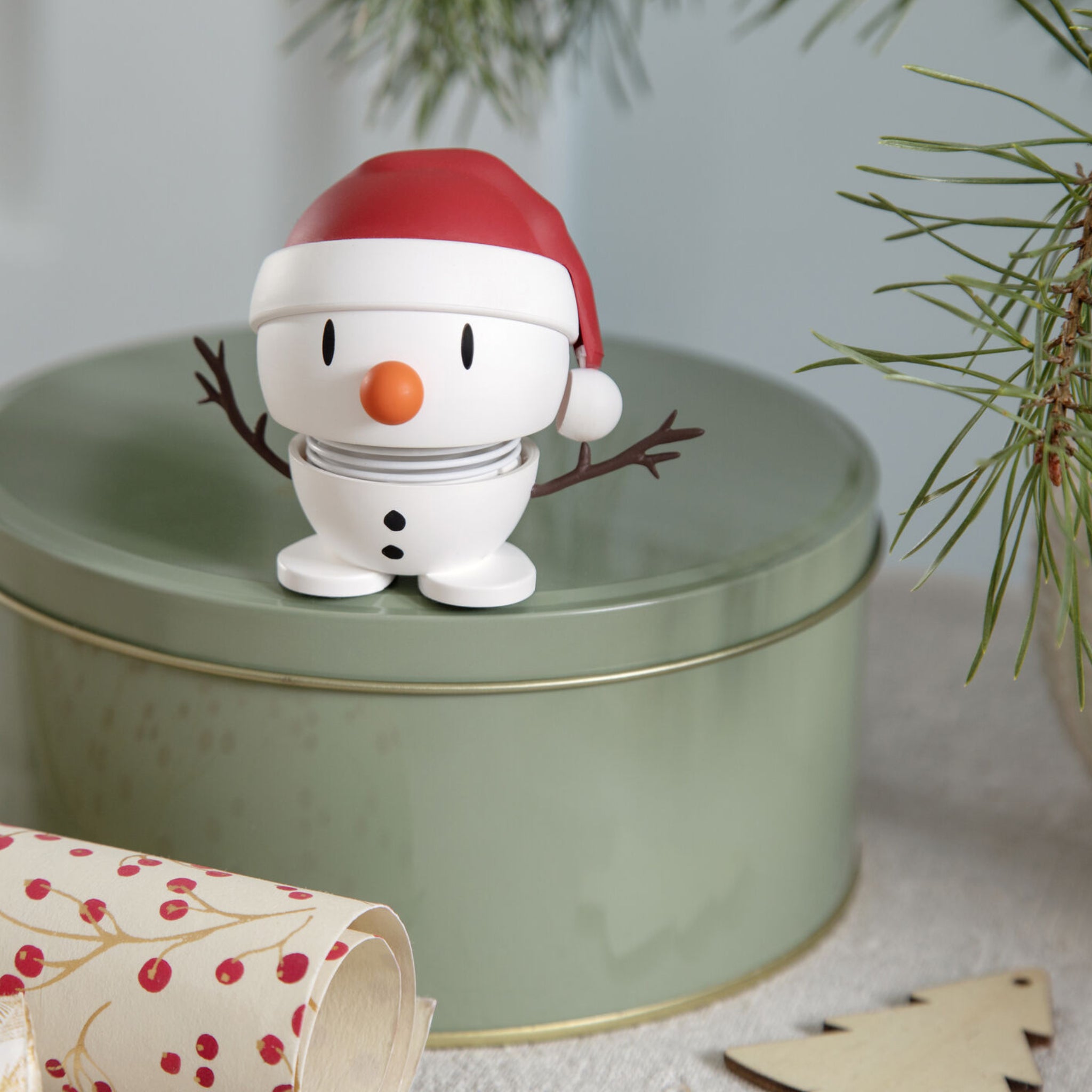 Het Hoptimist Zachte Kerstman Sneeuwman S Wit beeldje van Hoptimist, met een rode kerstmuts en twijgachtige armen, staat op een lichtgroen metalen blik te midden van dennentakken en feestelijke wikkels, en voegt vrolijke kerstdecoratie toe aan je kerstsfeer.