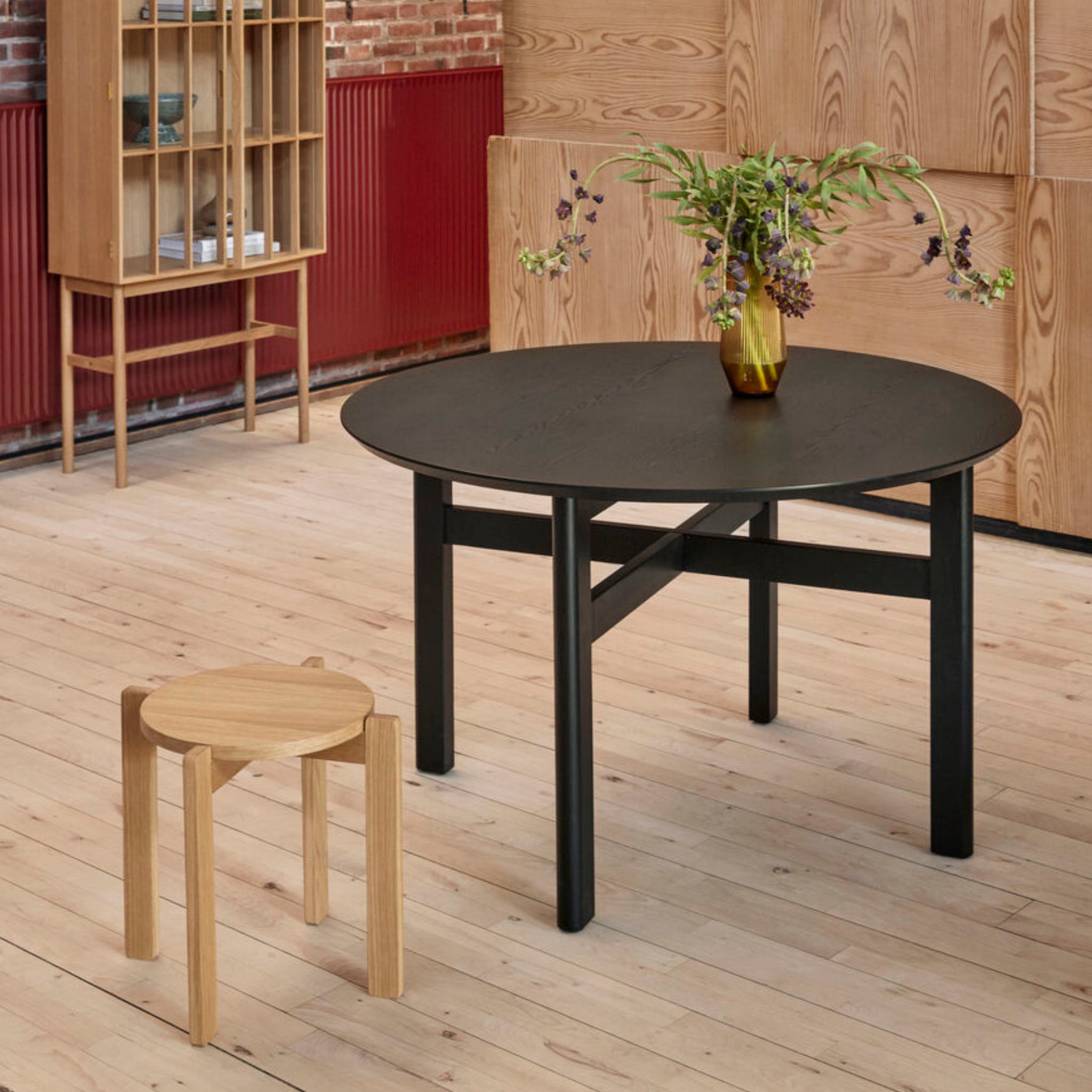 Een klein houten krukje staat naast de Hubsch Fjord Eettafel Essenhout Zwart D120, met daarop een vaas met groen, op een lichte houten vloer in een moderne kamer met houten wanden en een minimalistische inrichting.