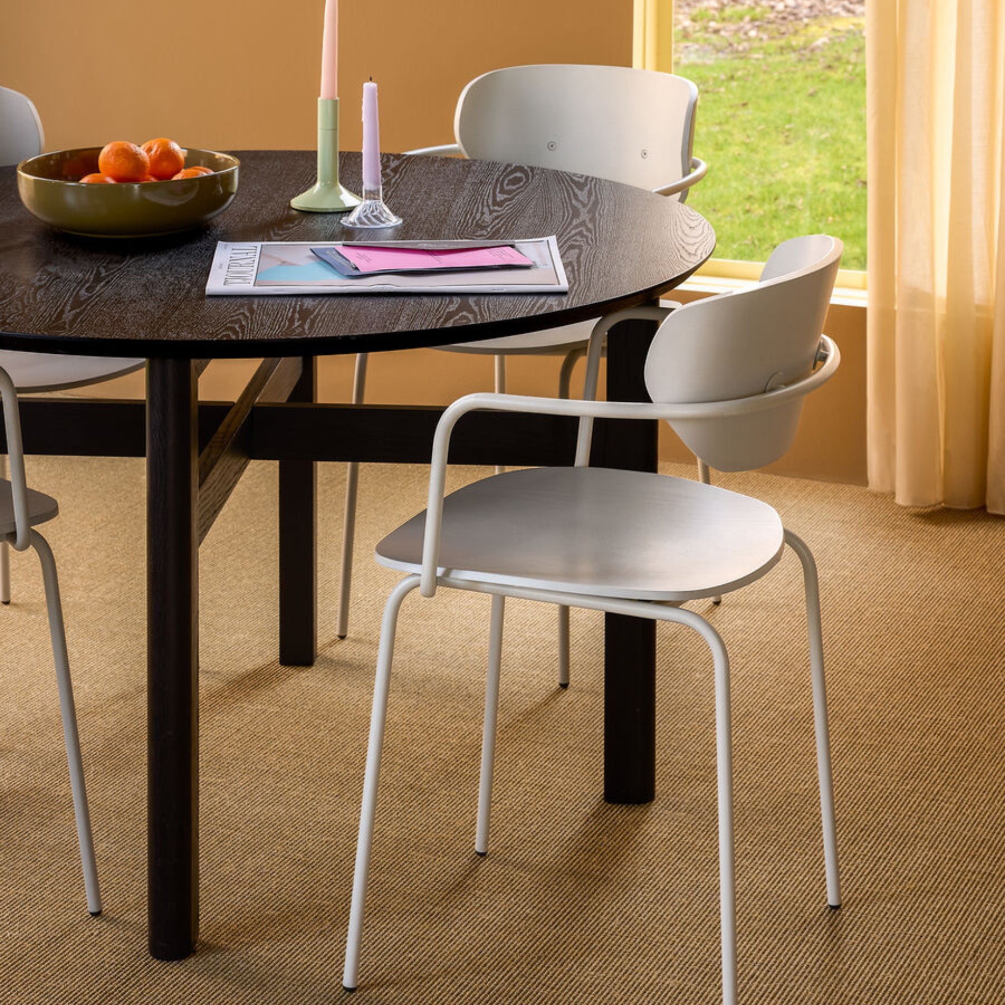 De Hubsch Fjord Eettafel Essenhout Zwart D120 van Hubsch, in combinatie met witte stoelen, een schaal sinaasappels, kaarsen en tijdschriften, staat op beige tapijt in een zonnige kamer met gele muren en crèmekleurige gordijnen.