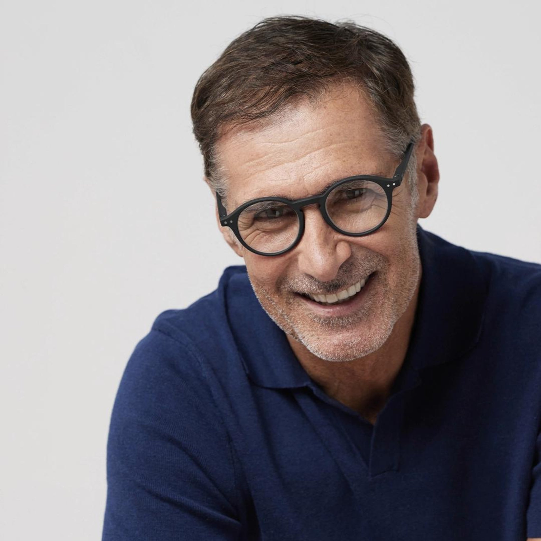 Een man van middelbare leeftijd met kort bruin haar en een getrimde baard glimlacht terwijl hij opzij kijkt, hij draagt Izipizi Leesbril #D Zwart van Izipizi en een marineblauwe trui, tegen een effen lichte achtergrond.