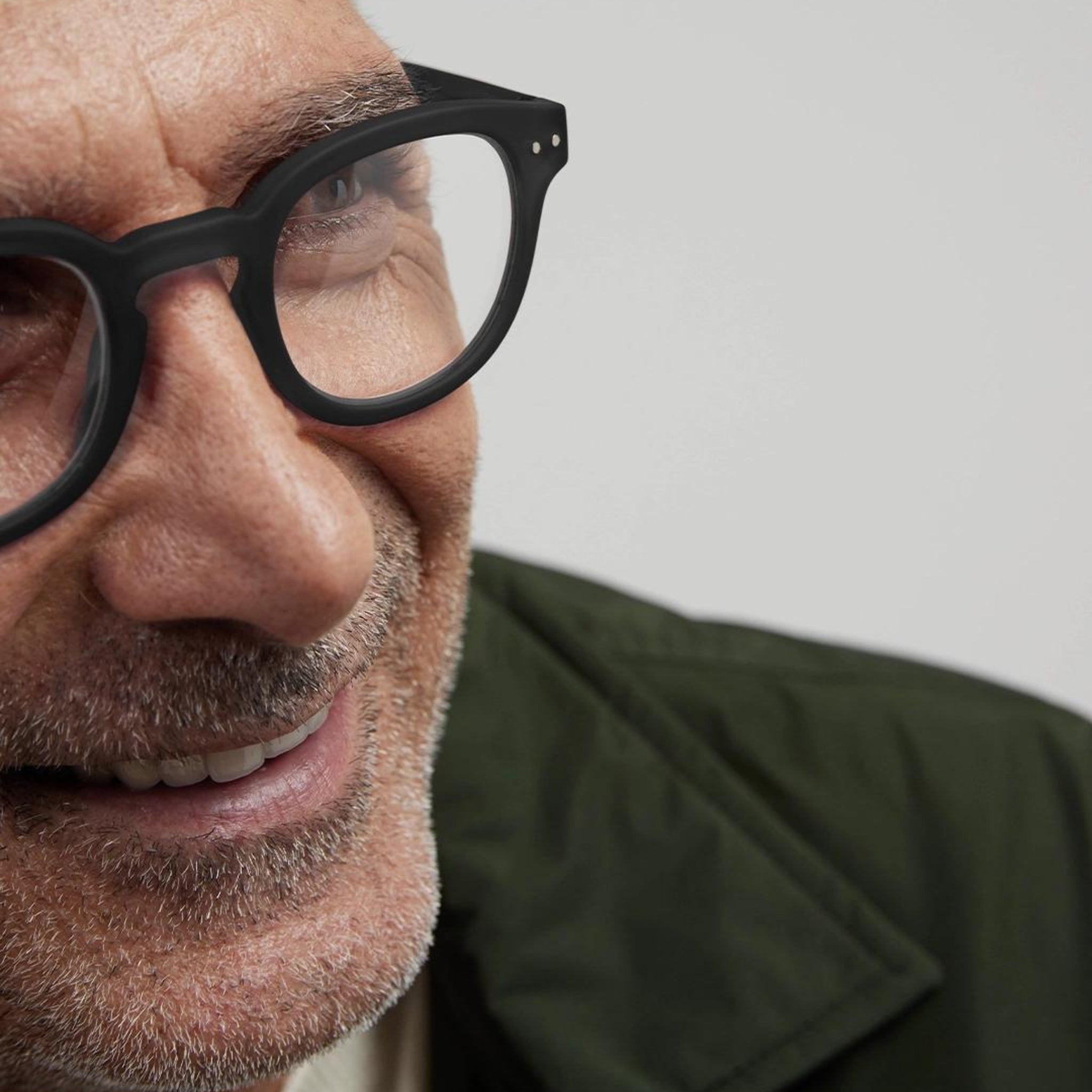 Close-up van een oudere man met een glimlach in een groene jas, die Izipizi Leesbril #C Zwart van Izipizi draagt. Alleen de linkerkant van zijn gezicht is te zien tegen een effen, lichte achtergrond.