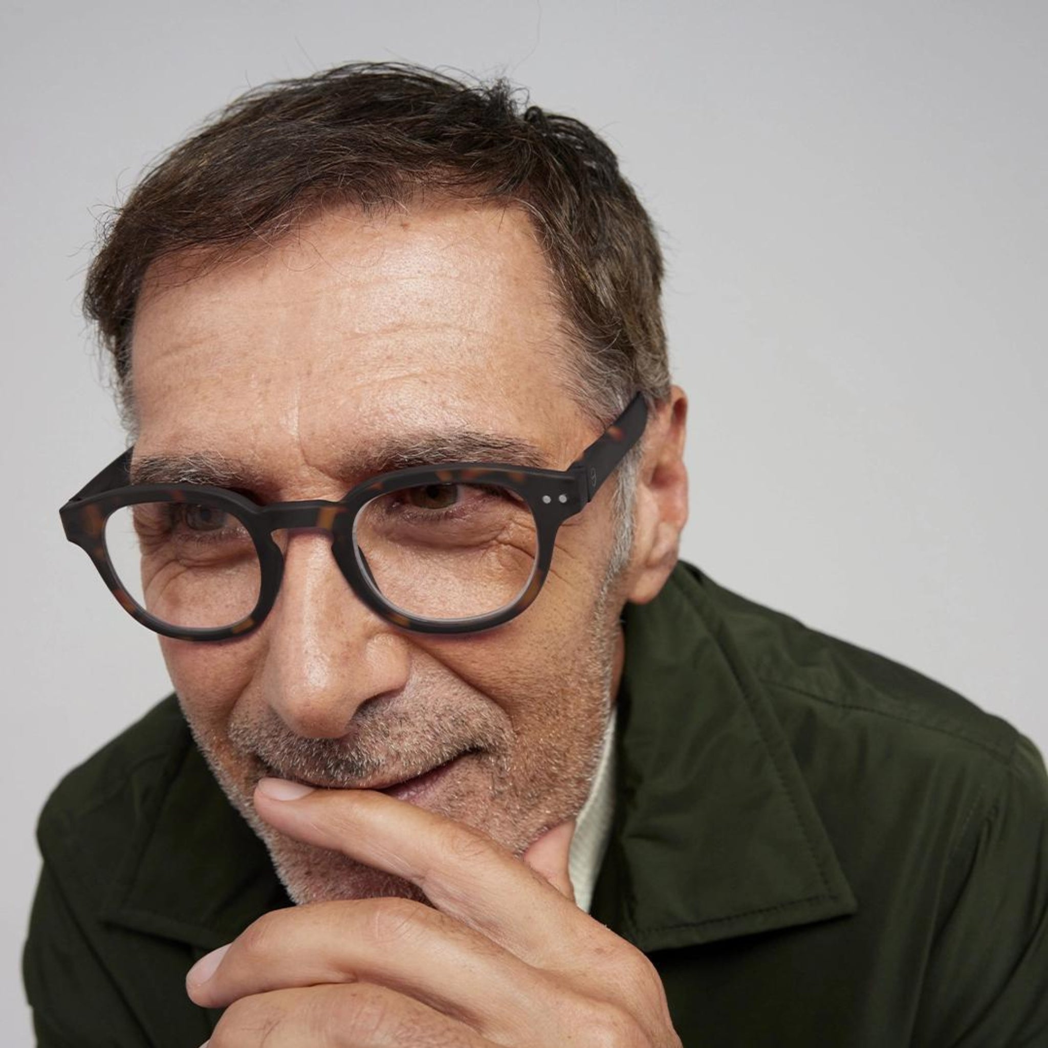 Een man van middelbare leeftijd met kort donker haar en een groen jasje kijkt peinzend naar de camera. Hij draagt een Izipizi Leesbril #C Tortoise van Izipizi. Zijn hand raakt zijn kin; de achtergrond is effen en lichtgekleurd.