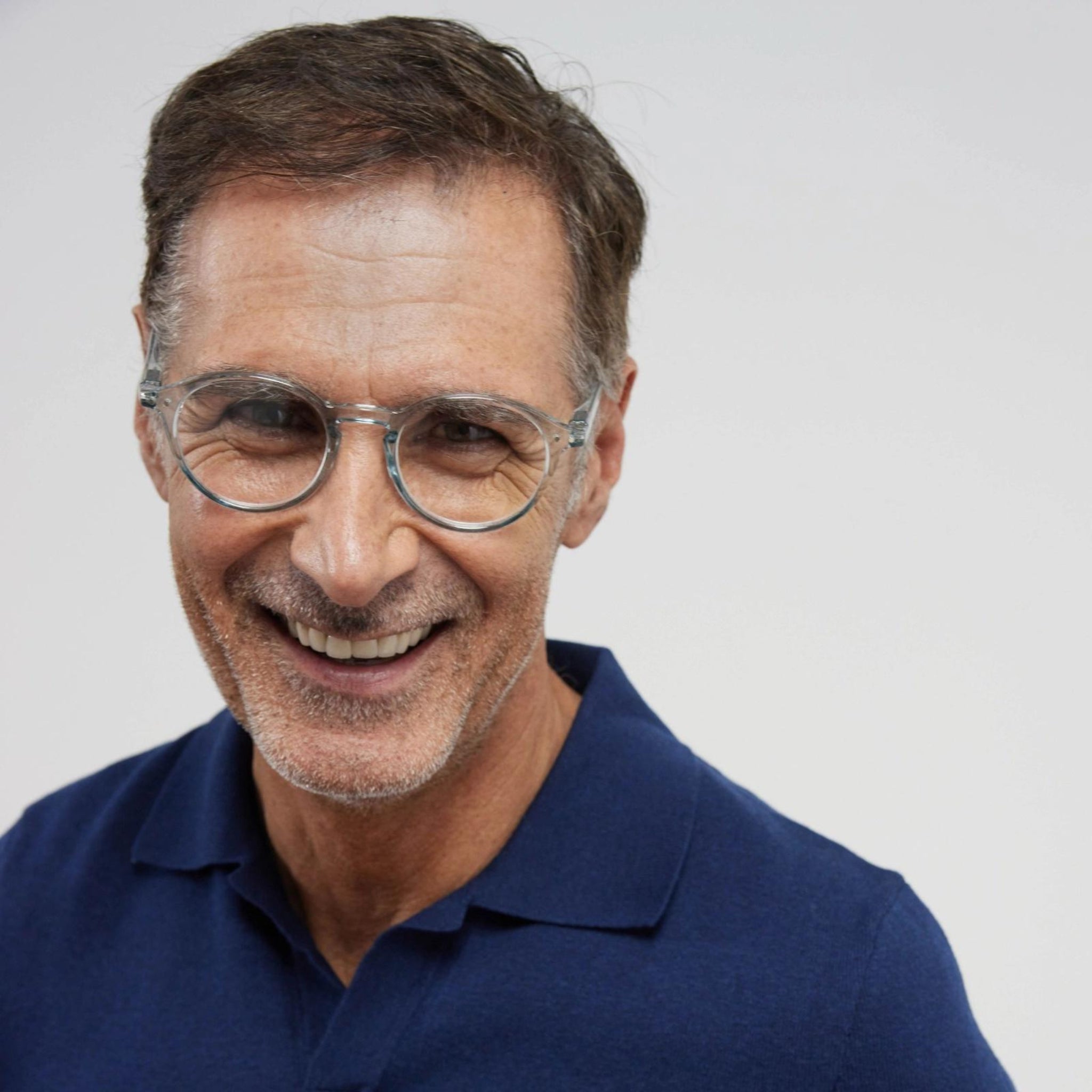 Een man van middelbare leeftijd met kort bruin haar, draagt een Izipizi Leesbril #D Frozen Blue met helder montuur en een marineblauwe polo, glimlacht hartelijk tegen een effen lichte achtergrond.