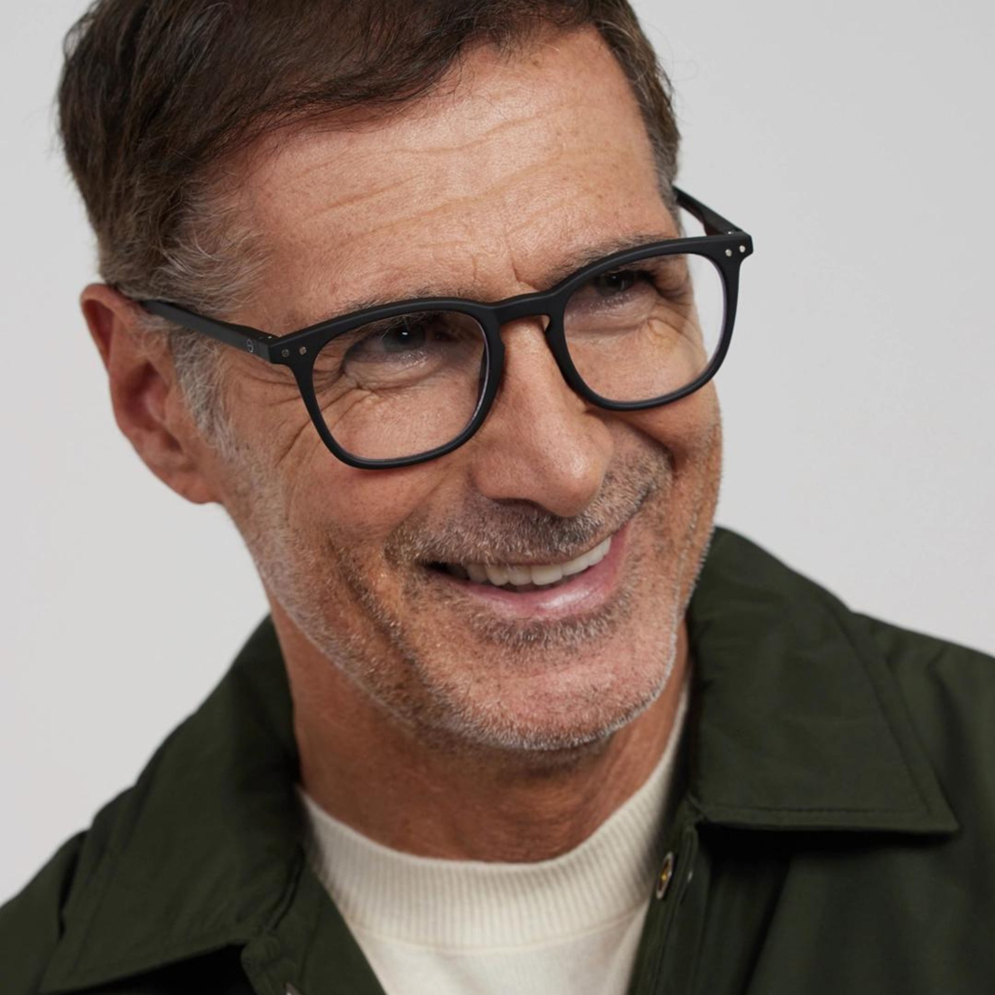 Een man van middelbare leeftijd met kort bruin haar, die een donkergroene jas, een crèmekleurige trui en de stijlvolle Izipizi Leesbril #E Zwart van Izipizi draagt, glimlacht terwijl hij iets opzij kijkt tegen een effen lichte achtergrond.