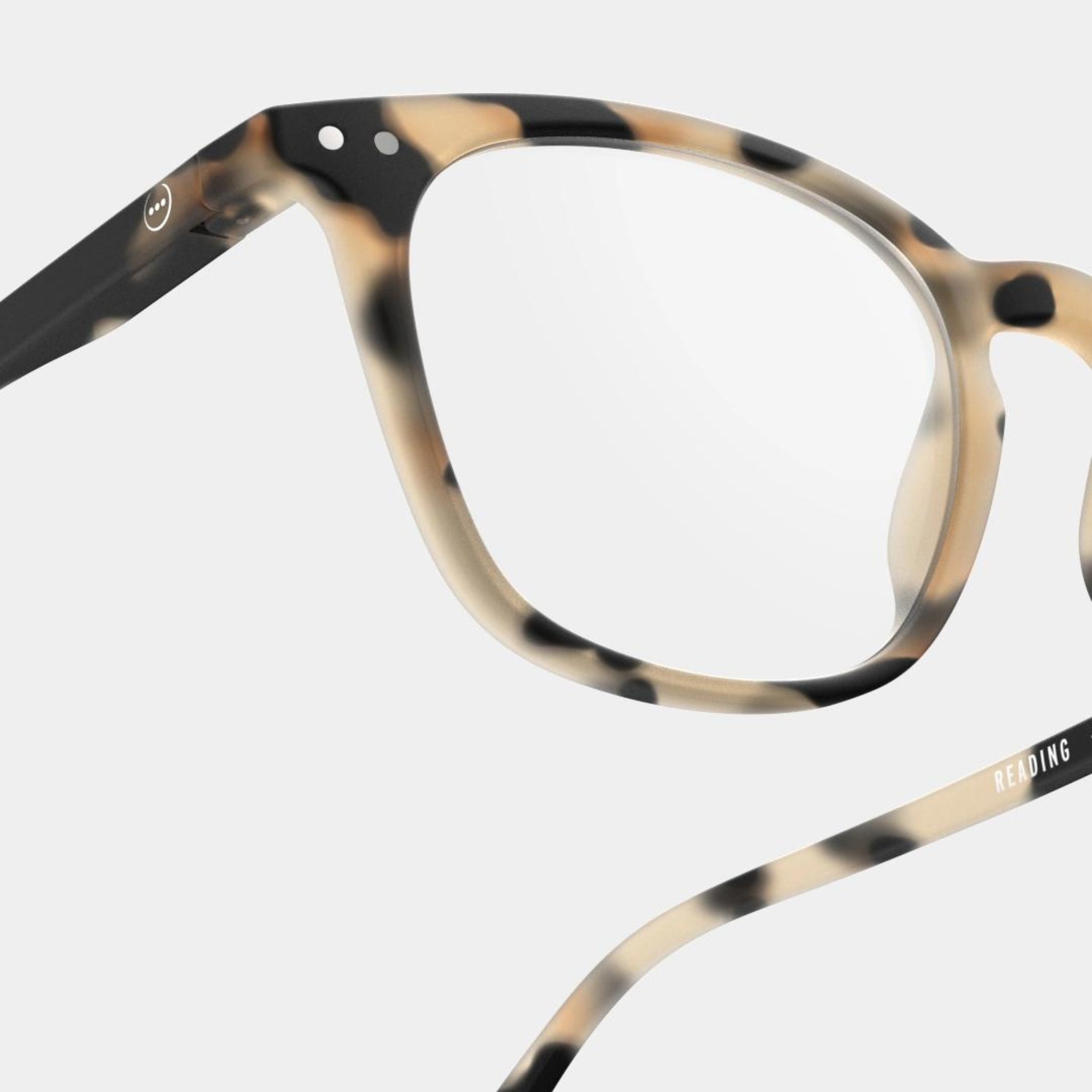 Close-up van Izipizi Leesbril #E Light Tortoise van Izipizi, een stijlvolle unisex leesbril met heldere glazen. Het schildpadmontuur in beige, bruine en zwarte tinten heeft "READING" op de arm en is geschikt voor zowel mannen als vrouwen.