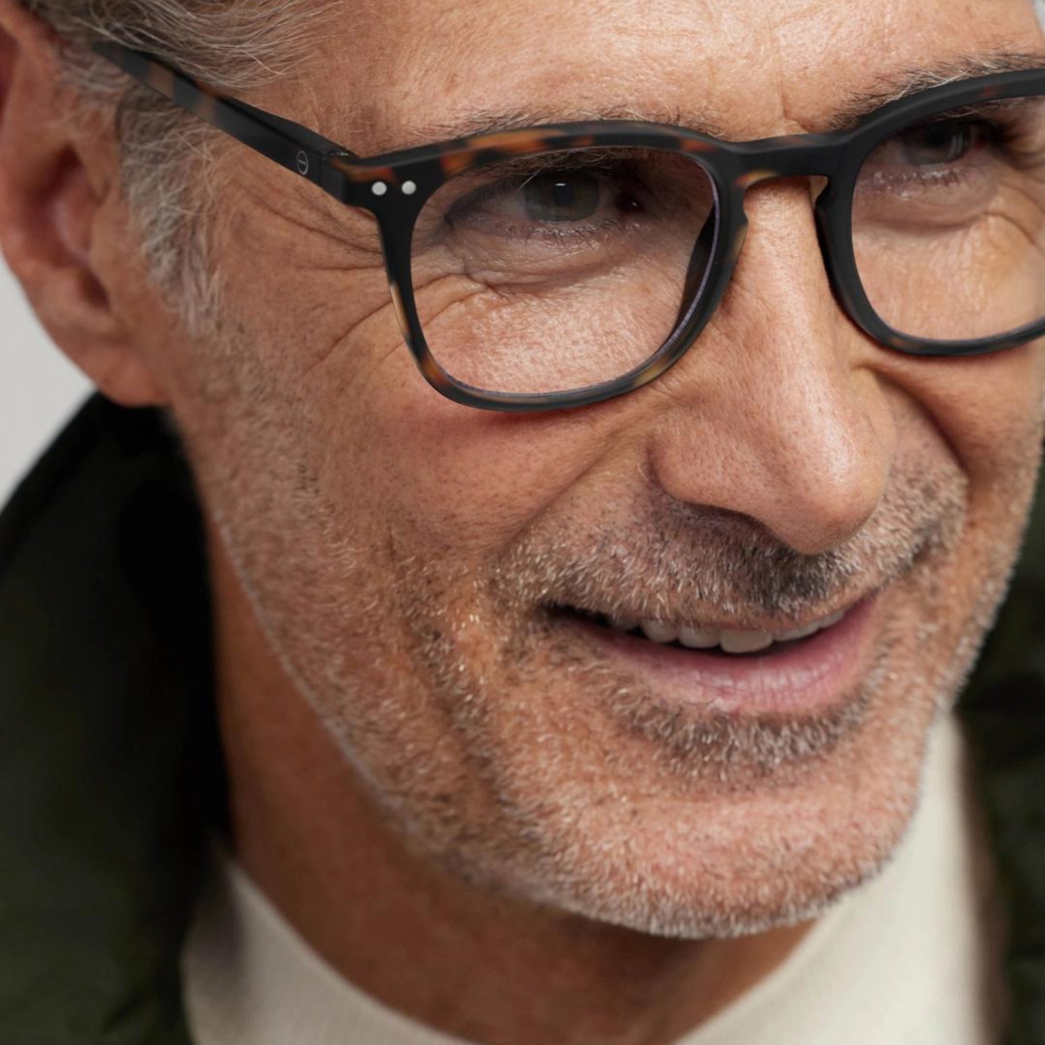 Close-up van een lachende man van middelbare leeftijd met kort grijs haar en stoppelbaard, die een Izipizi Leesbril #E Tortoise van Izipizi draagt met een bril met zwarte montuur, een wit overhemd en een donker jasje voor een stijlvolle look.