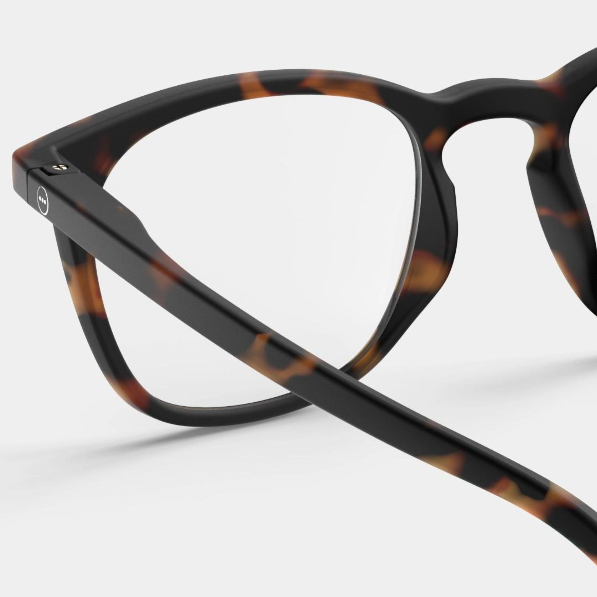 Close-up van de bovenste helft van de Izipizi Leesbril #E Tortoise van Izipizi, met een montuur in zwart en bruin schildpadmotief met zichtbare scharnier en veerarm tegen een effen lichte achtergrond. Een modieuze leesbril voor elke stijl.