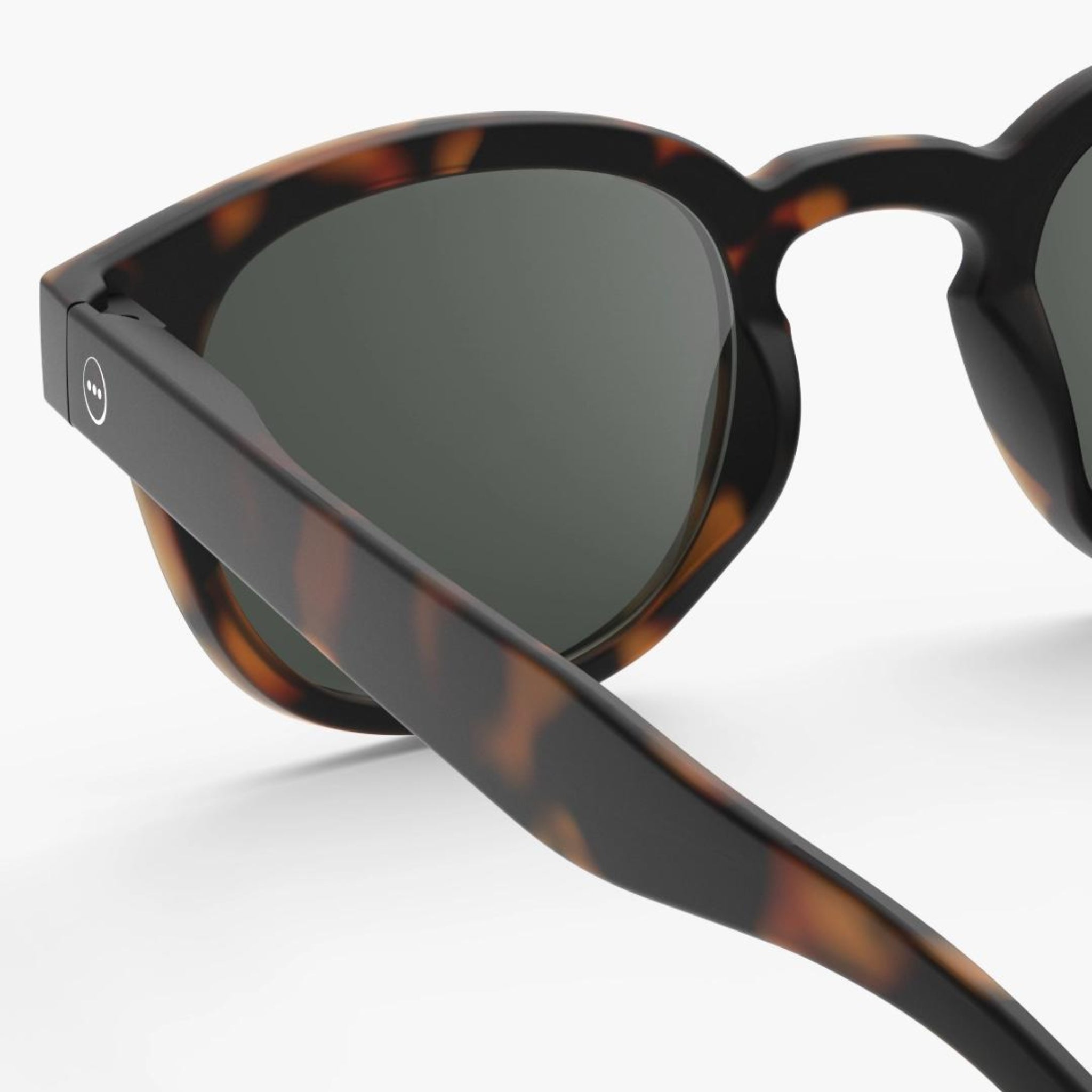 Close-up van de Izipizi Zonnebril Sterkte #C Tortoise van Izipizi, met donkere glazen en gedetailleerde scharnieren op een glad, mat schildpadmontuur. De effen witte achtergrond benadrukt het stijlvolle unisex UV-beschermende ontwerp.