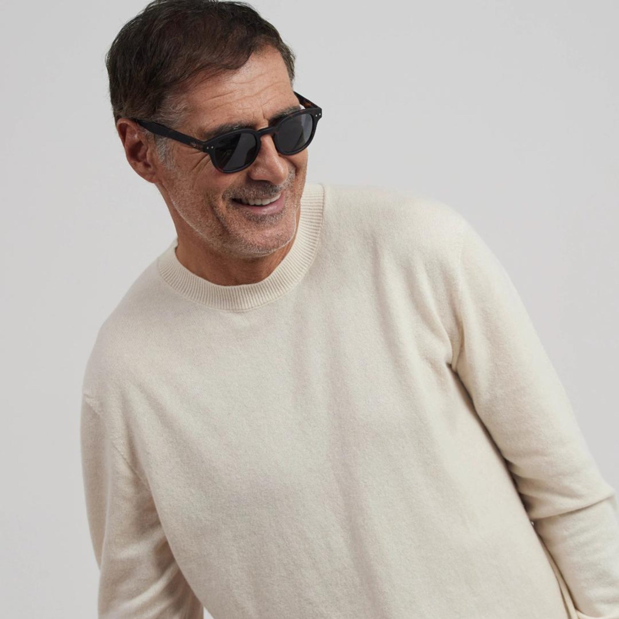 Een lachende man van middelbare leeftijd met kort donker haar en een getrimde baard draagt de Izipizi Zonnebril #C Tortoise van Izipizi en een crèmekleurige trui, staande tegen een effen lichte achtergrond.