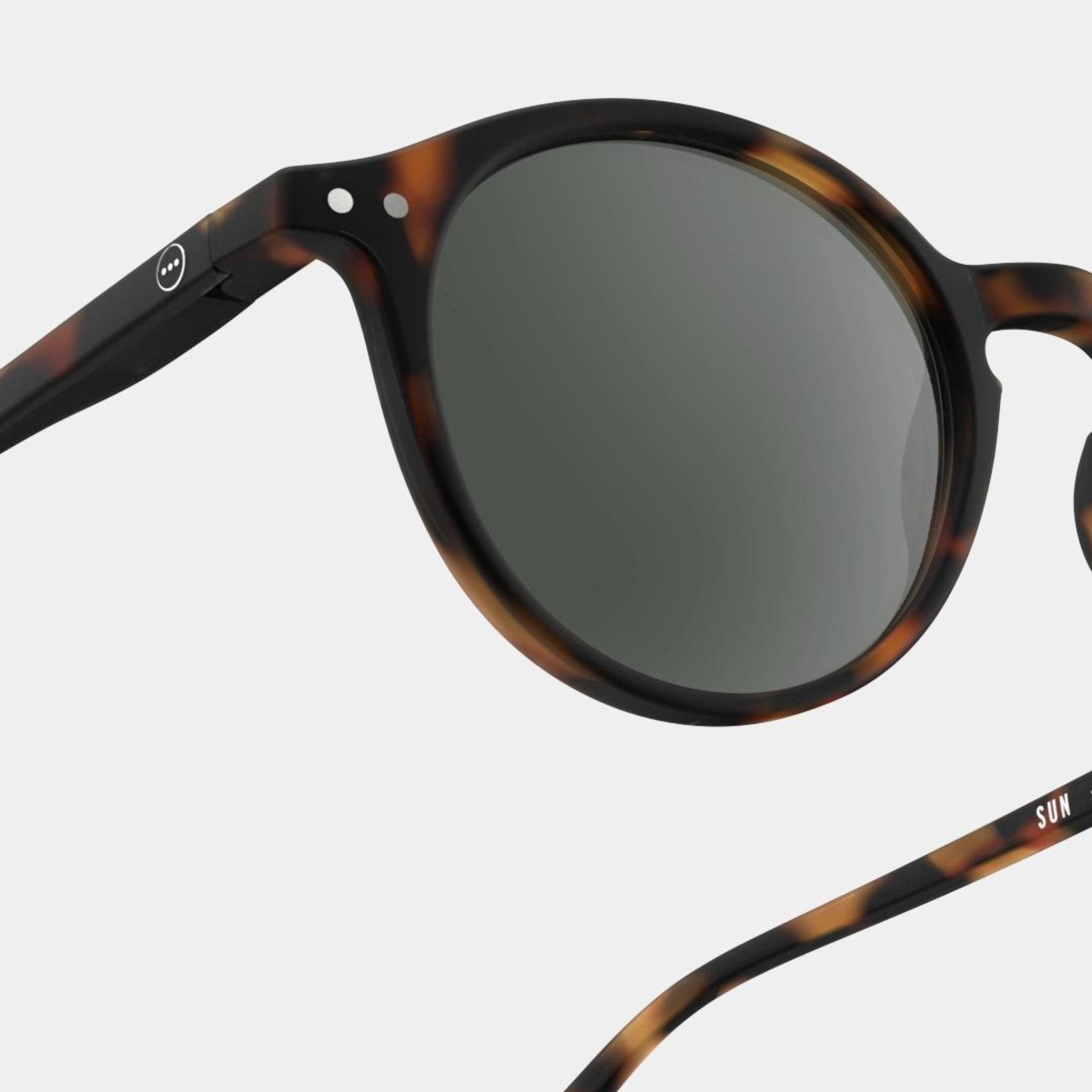 Close-up van de Izipizi Zonnebril #D Tortoise van Izipizi, met een schildpadmontuur, ronde donkere glazen, subtiel logo op de arm en UV-bescherming. Weergegeven op een lichtgrijze achtergrond voor stijlvolle veiligheid tegen de zon.