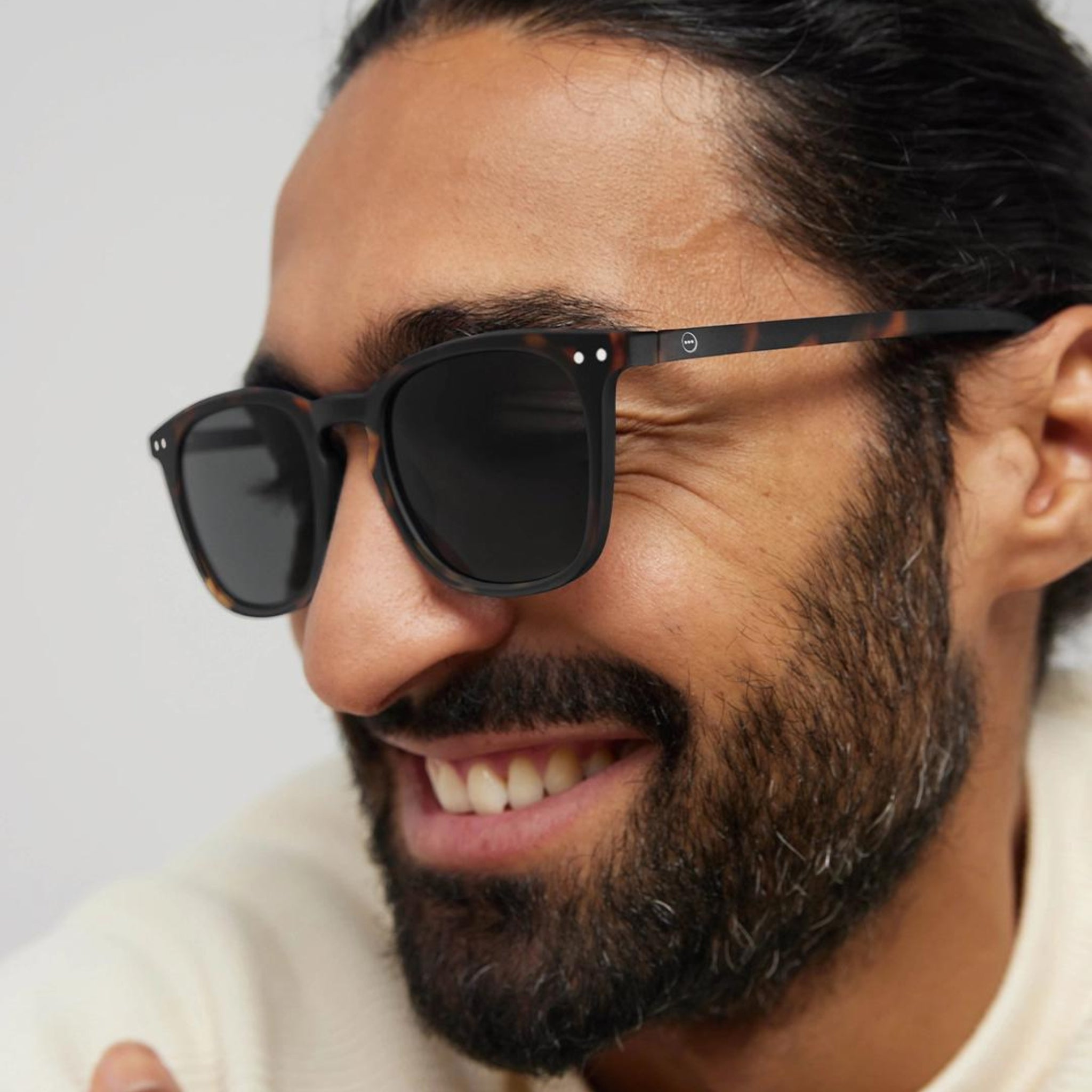 Een man met lang donker haar en een baard glimlacht terwijl hij de Izipizi Zonnebril #E Tortoise draagt. Zijn stijlvolle zonnebril van Izipizi steekt af tegen zijn lichte trui en de effen achtergrond, waardoor de focus op zijn look blijft liggen.