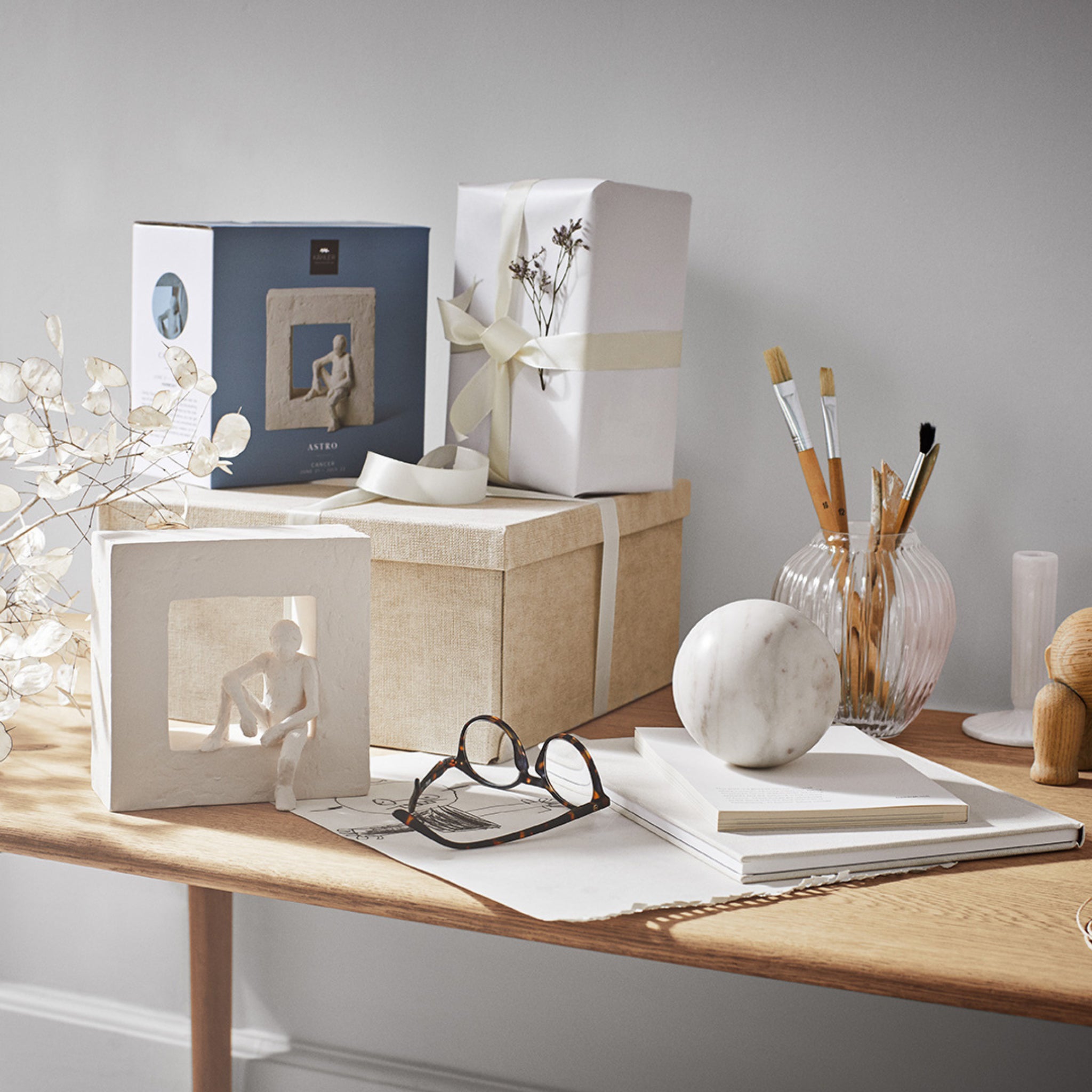 Een houten bureau met kunstbenodigdheden, een notitieblok, een bril, een Kähler vaas met kwasten, een ingelijste Kähler Astro Sterrenbeeld Sculptuur Kreeft, een marmeren bol, twee ingepakte cadeaus en droogbloemen tegen een neutrale muur.