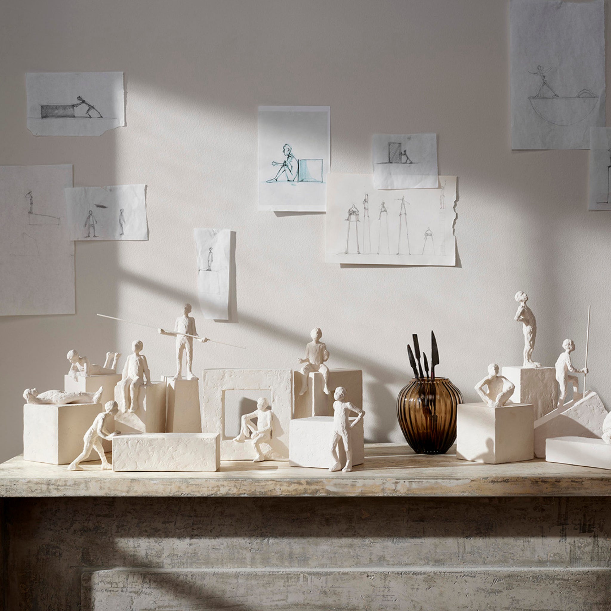 Kähler Astro Sterrenbeeld Sculptuur Kreeft van Kähler staat tentoongesteld op een rustieke schouw, met schetsen van soortgelijke figuren erachter geplakt. Zonlicht accentueert de witte mensfiguur van klei en creëert een vredige sfeer.