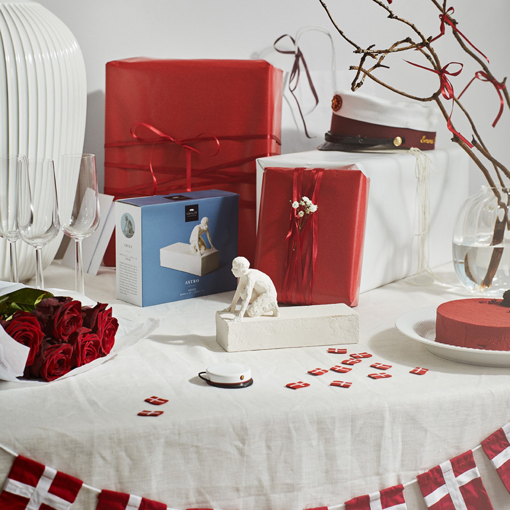 Een gedekte tafel voor een feest met ingepakte cadeaus, een Deense afstudeerpet, rode rozen, champagneglazen, een rode taart, kleine Deense vlaggetjes en de Kähler Astro Sterrenbeeld Sculptuur Ram van Kähler. Rode en witte decoraties accentueren het feestelijke tafereel.