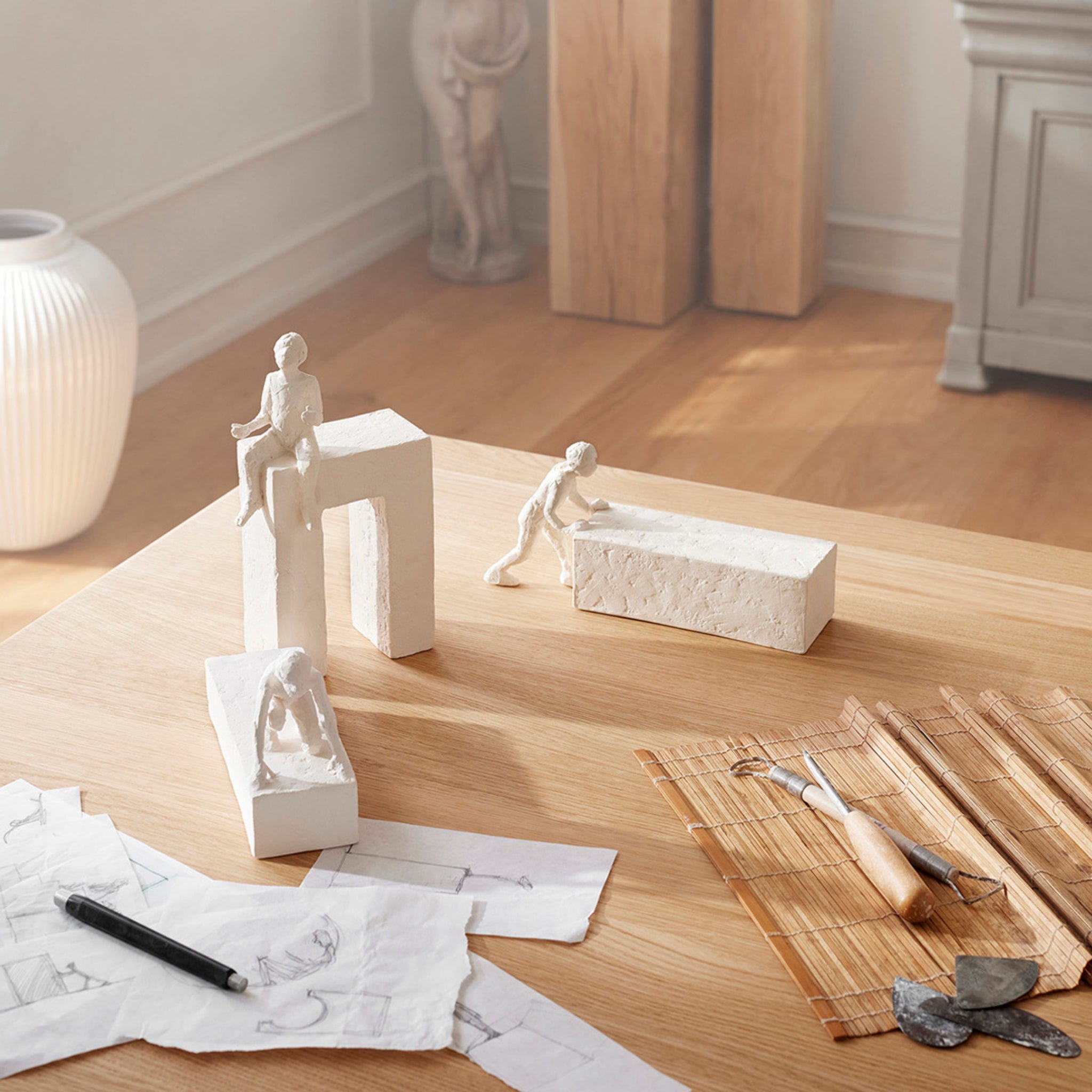 Op een houten tafel staat een kleine witte Kähler Astro Sterrenbeeld Sculptuur Ram van Kähler, samen met teken- en beeldhouwgereedschap, schetsen op papier en een bamboemat. Zonlicht stroomt naar binnen door een raam, waardoor de creatieve werkruimte helderder wordt.
