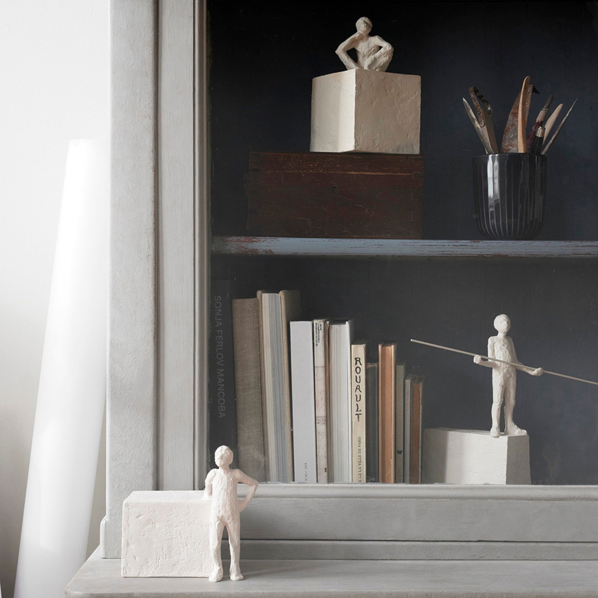 Op een kastplank staan boeken, een beker verfkwasten en drie kleine Kähler Astro Sterrenbeeld Sculptuur Schorpioen steengoede sculpturen van menselijke figuren - één bedacht op een kubus, één met een paal en één naast een rechthoekig blok.