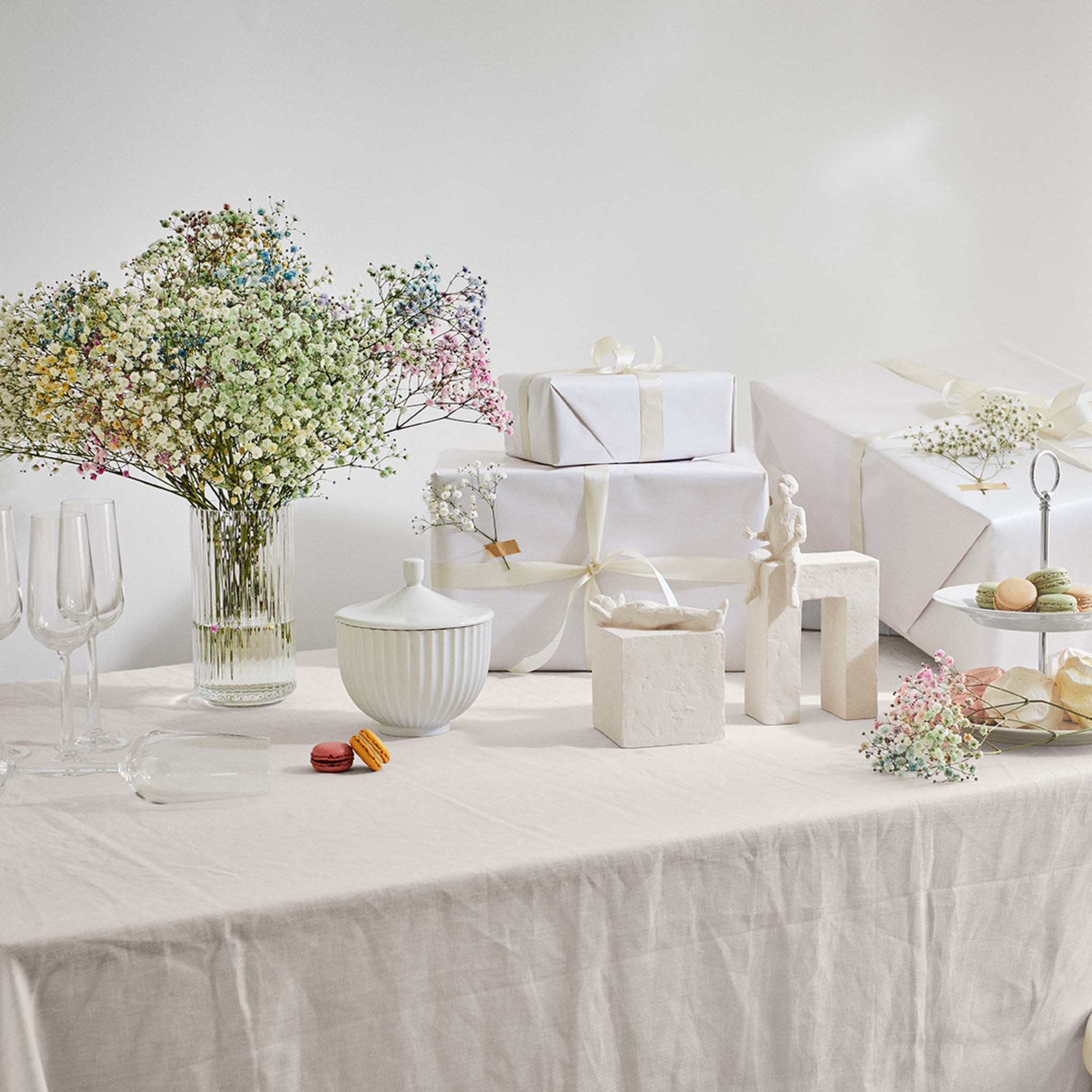 Een gedekte tafel met een wit tafelkleed, een vaas met gipskruidbloemen, ingepakte cadeautjes, wijnglazen, macarons, Kähler Astro Sterrenbeeld Sculptuur Tweeling van Kähler keramiek en een trapsgewijs dienblad creëert een elegante feestelijke sfeer.