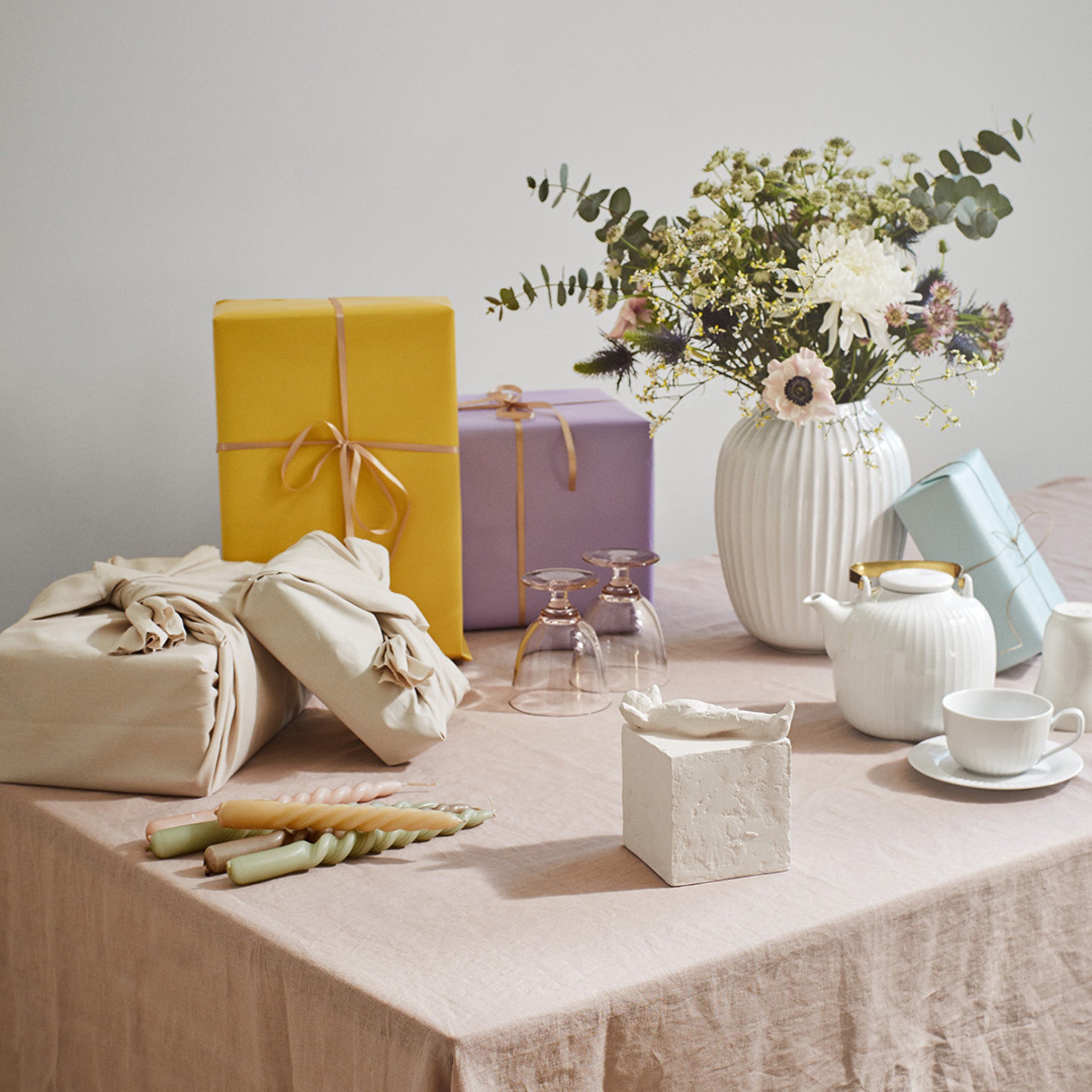Een gedekte tafel met ingepakte cadeautjes, een witte vaas met bloemen, een theepot, theekopje, glazen, gedraaide kaarsen en de Kähler Astro Sterrenbeeld Sculptuur Vissen van Kähler, allemaal gerangschikt op een beige tafelkleed.