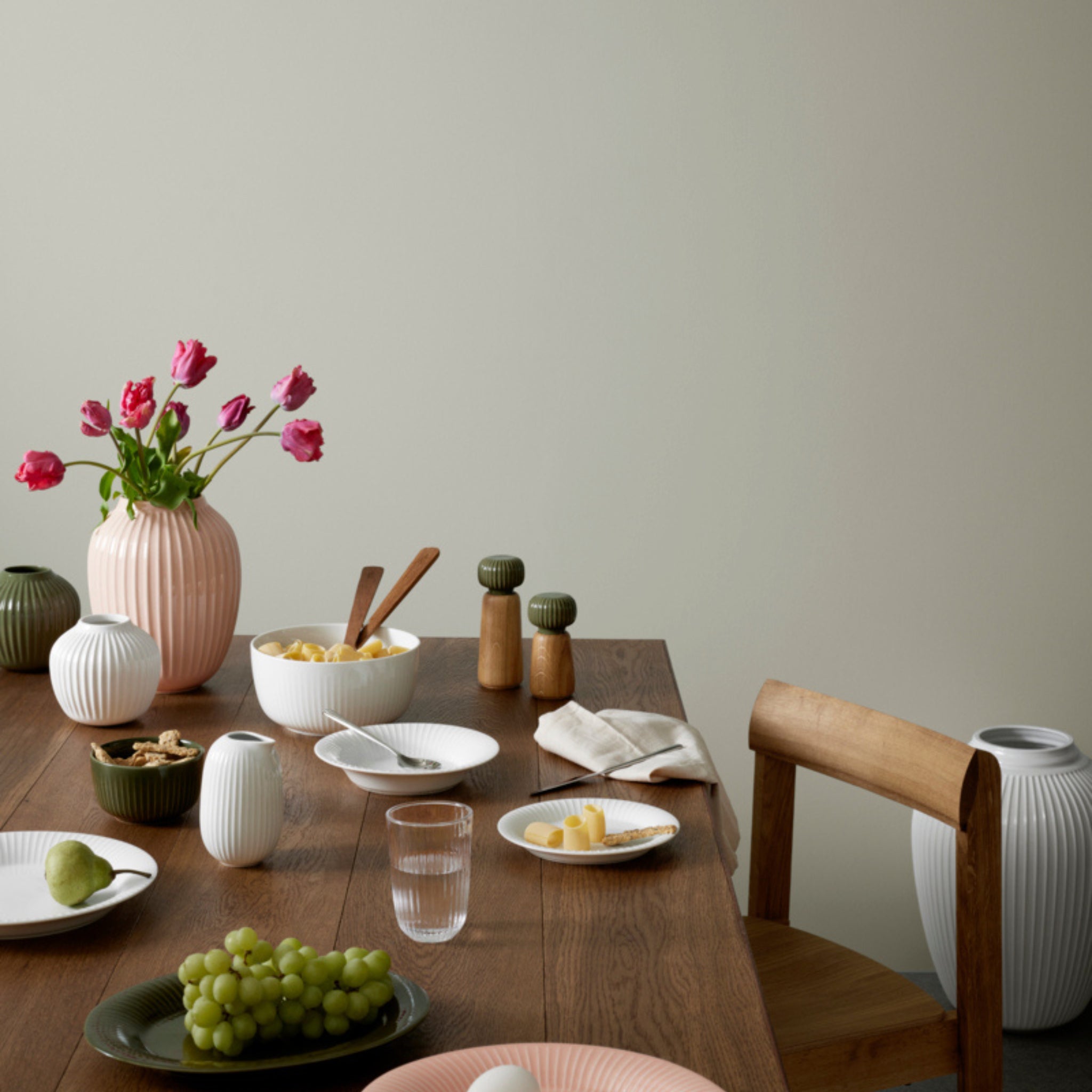 Een houten eettafel is gedekt met borden druiven en peren, schaaltjes eten, een glas water, een Kähler Hammershoi Kruidenmolen Donkergroen S, peper- en zoutstel, roze tulpen in vazen en een houten stoel ernaast.