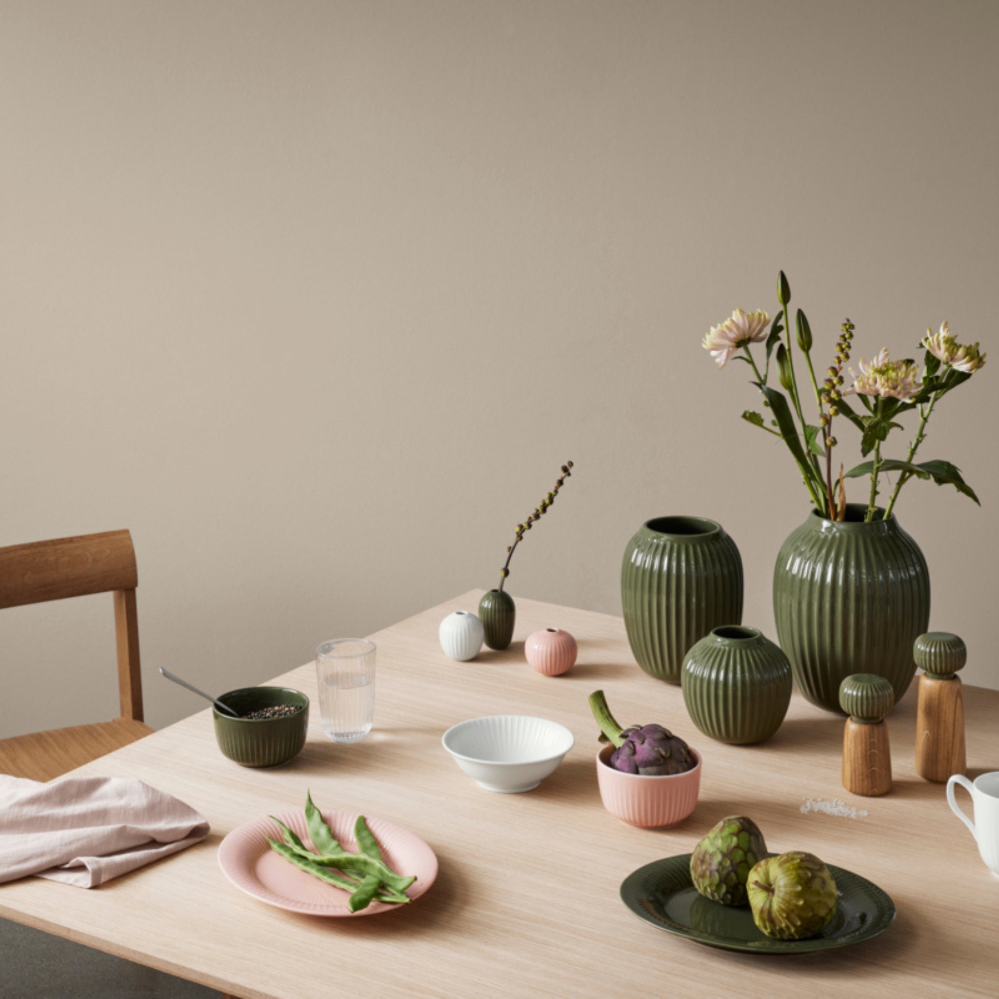 Een houten tafel gedekt met groen en roze keramiek van Kähler Hammershøi, verse bloemen, een glas water, een servet, artisjokken en de Kähler Hammershoi Kruidenmolen Donkergroen S staat tegen een beige muur in een minimale, stijlvolle opstelling.