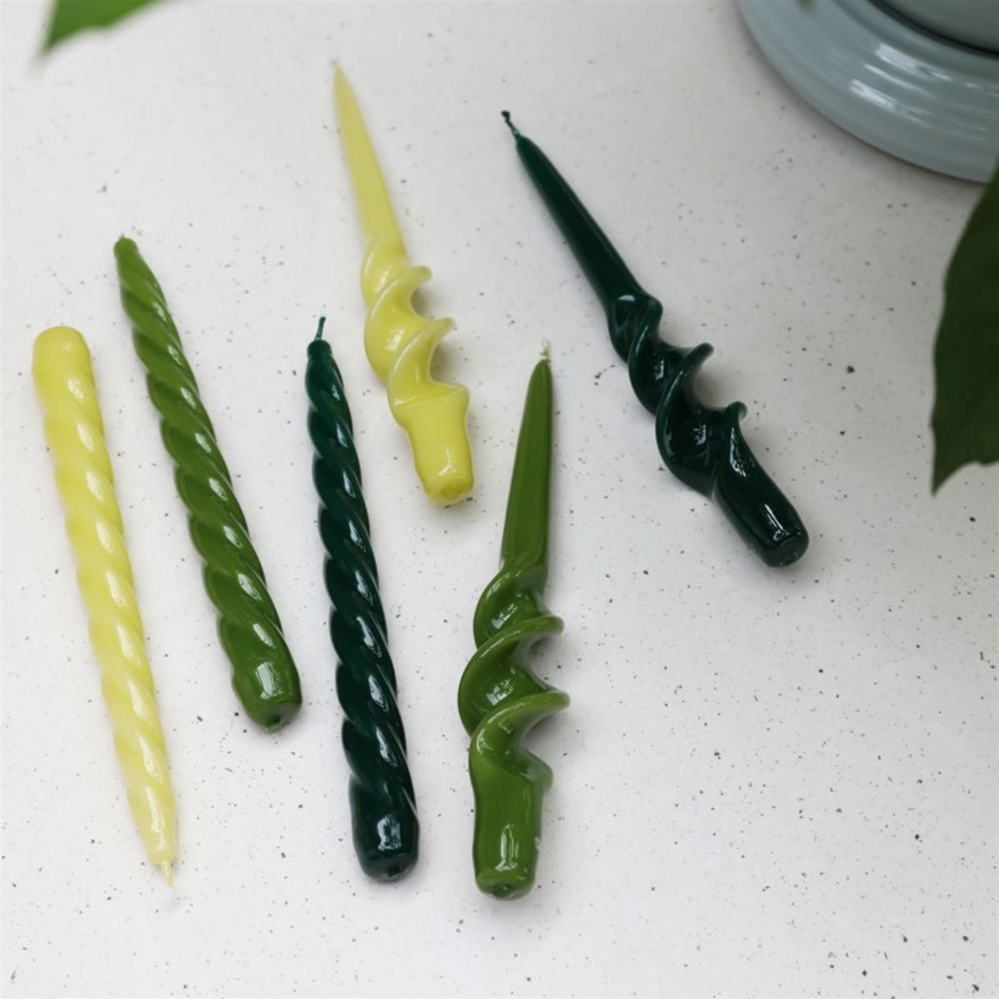 Drie &Klevering Bravo Kaarsen Groen gedraaide kaarsen staan op een wit gespikkeld oppervlak, geaccentueerd door groene bladeren aan de randen.