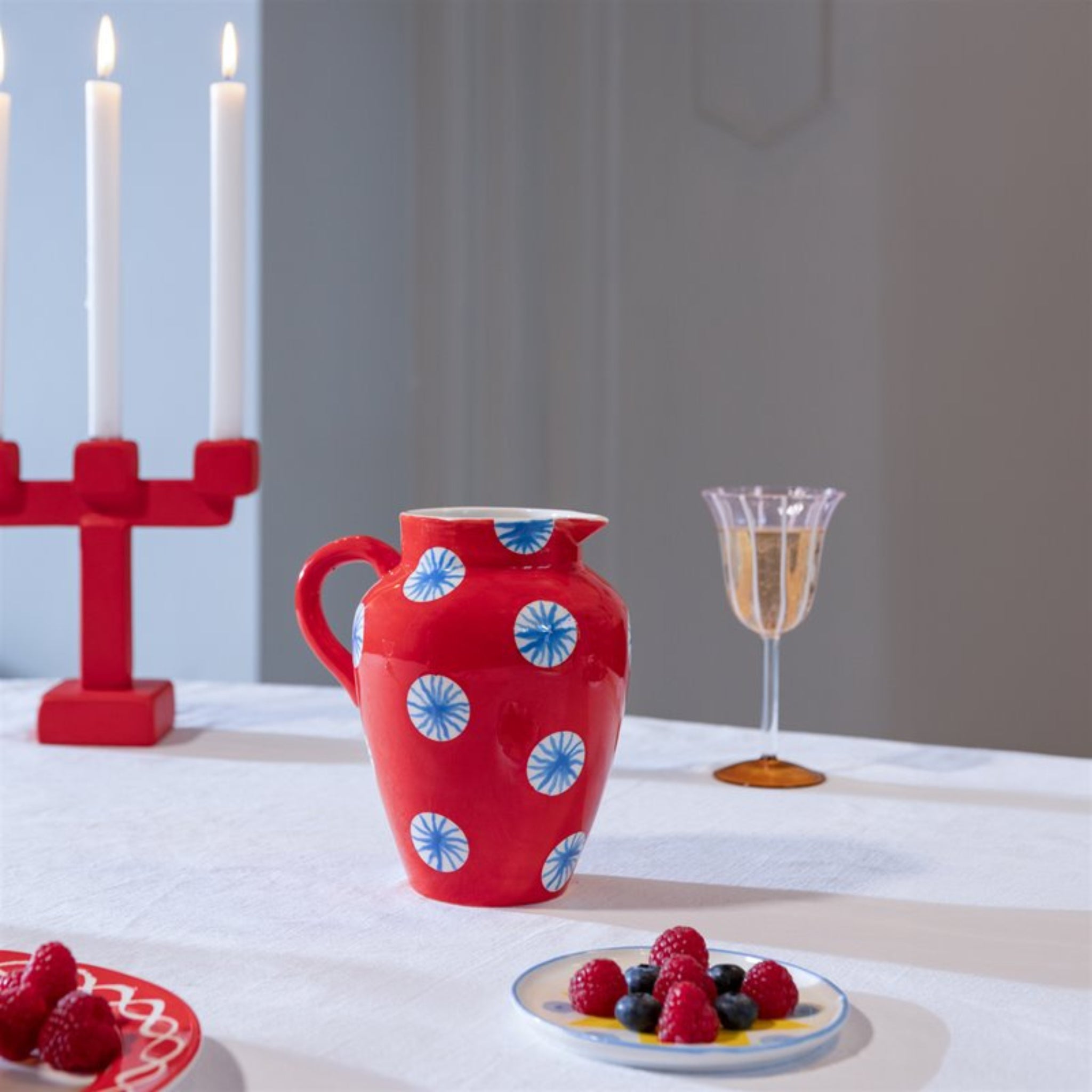Een &Klevering Festivo Kan Rood 1,5L met blauwe en witte cirkels rust op een wit tafelkleed naast een bord bessen, een champagneglas en een rode kandelaar met drie brandende kaarsen, die samen een charmante decoratieve display vormen.